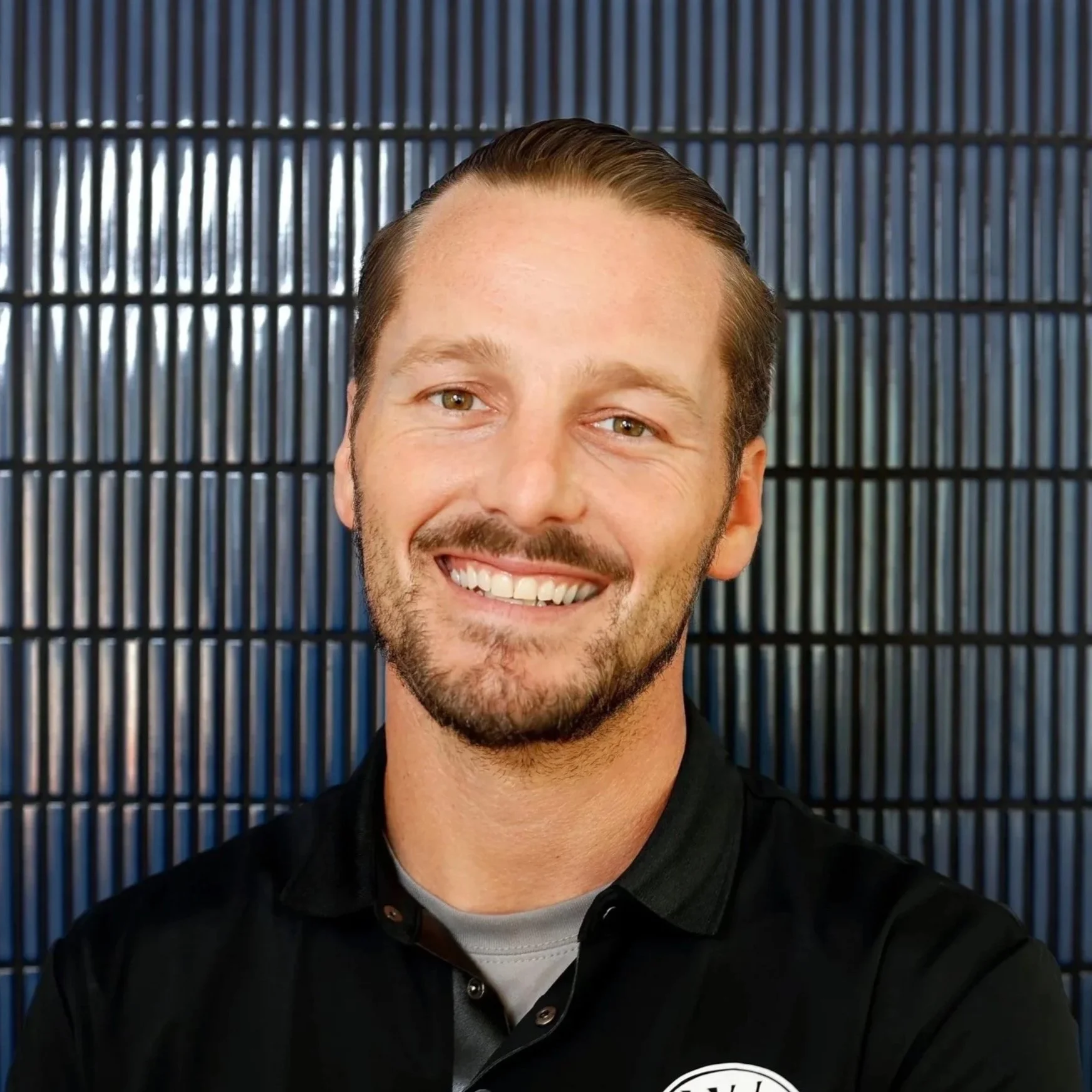 A smiling man with short light brown hair and a beard, wearing a black collared shirt, standing in front of a dark blue grid-patterned wall.