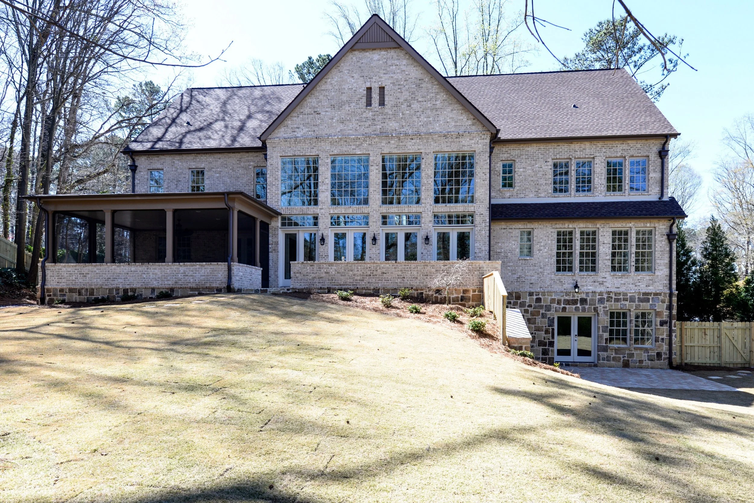 Rear view of a large brick house with multiple windows, a covered patio, and a landscaped yard with a slight slope.