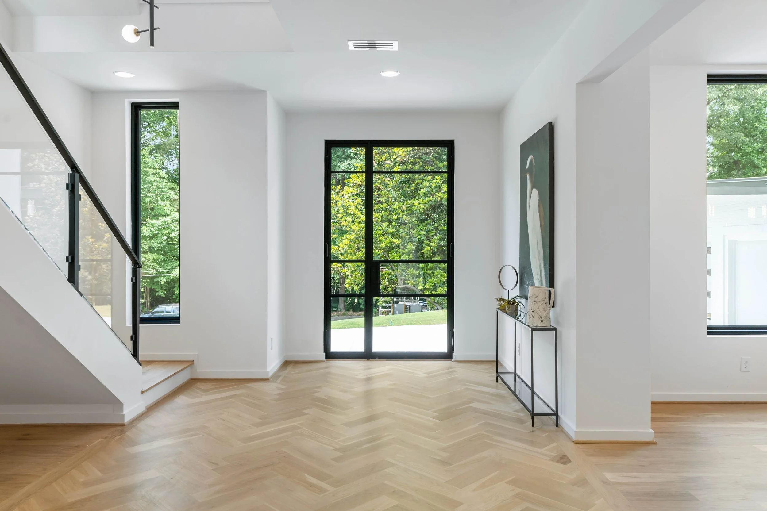 Bright, modern living room with white walls, light wooden herringbone floor, large black-framed window, and a glass sliding door leading outside. Decor includes a minimalist console table with artwork and decorative items.