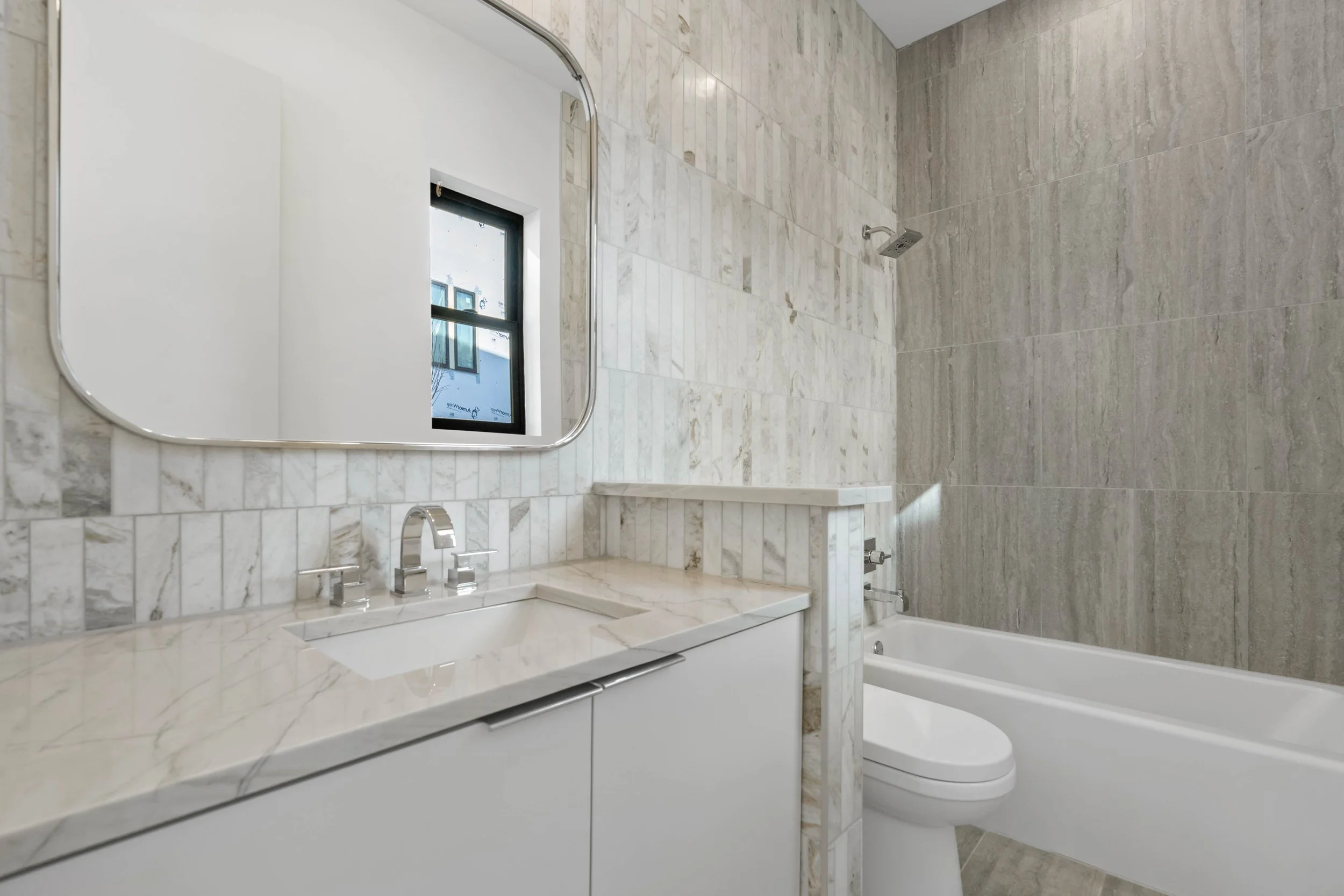 Modern bathroom with marble countertops, a sink, a wall-mounted mirror, a window, a toilet, and a bathtub with a showerhead.