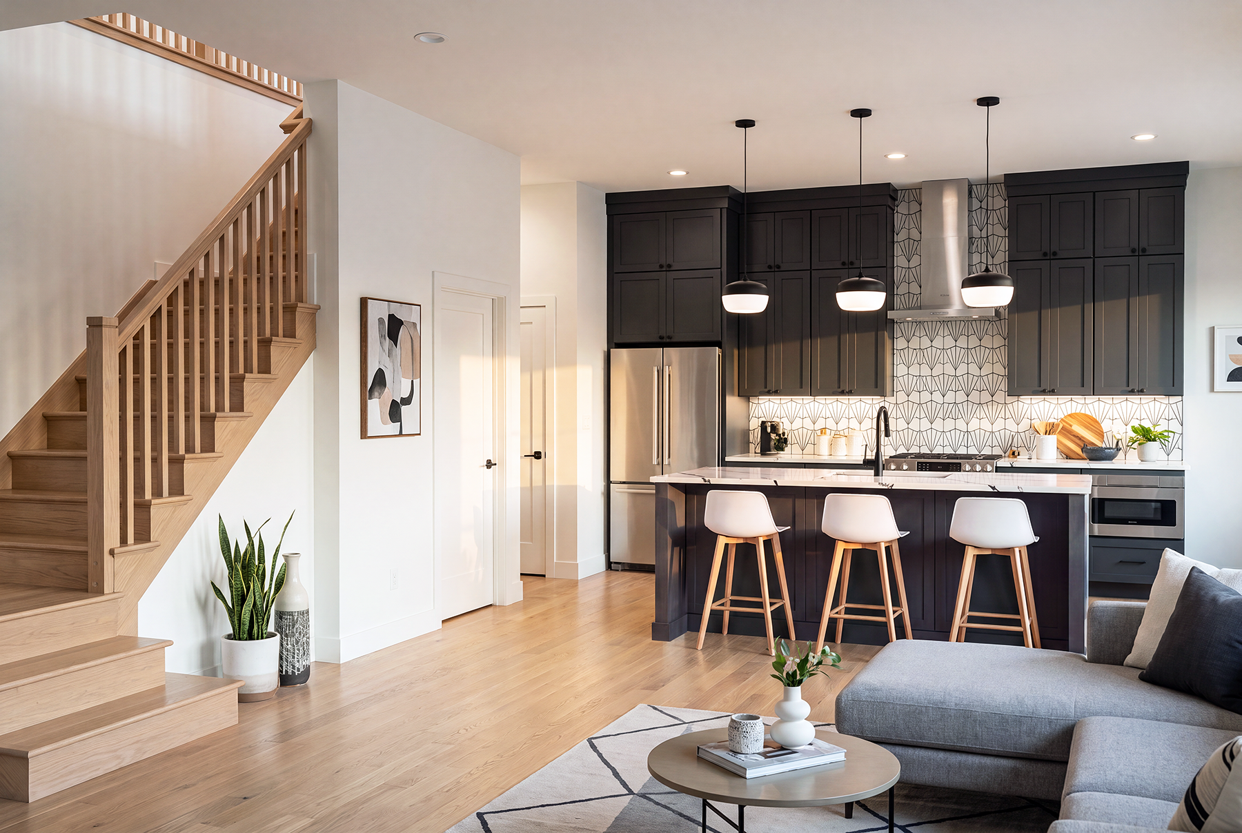 Modern kitchen with dark cabinets, white countertops, a patterned backsplash, and a kitchen island with three pendant lights and four bar stools. Adjacent living space with a gray sectional sofa, coffee table, and decorative plants. Light hardwood fl