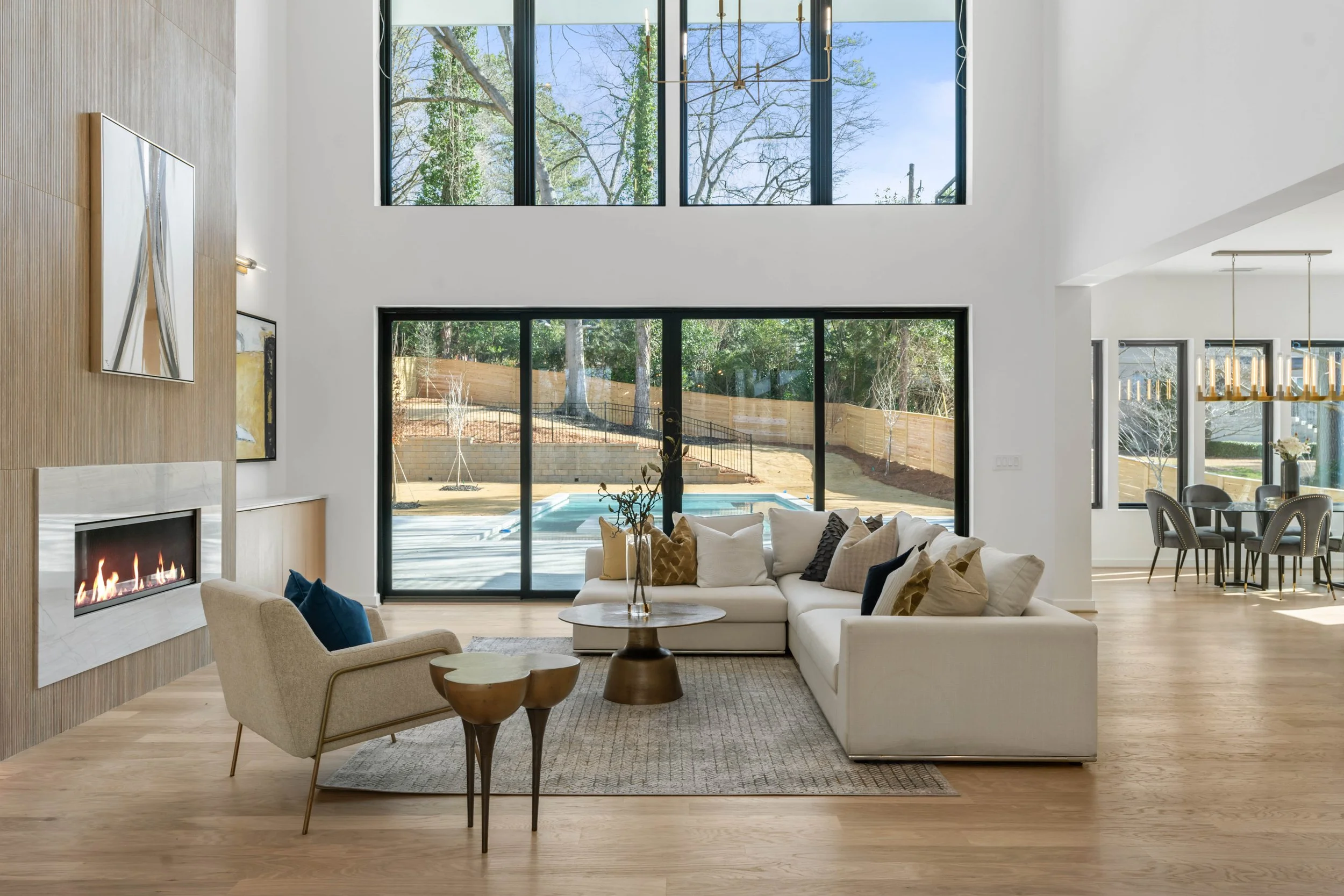 Spacious modern living room with large floor-to-ceiling windows showing a backyard with a pool, dining area in the background, and minimalist furniture including a white sofa, beige armchair, and a fireplace.