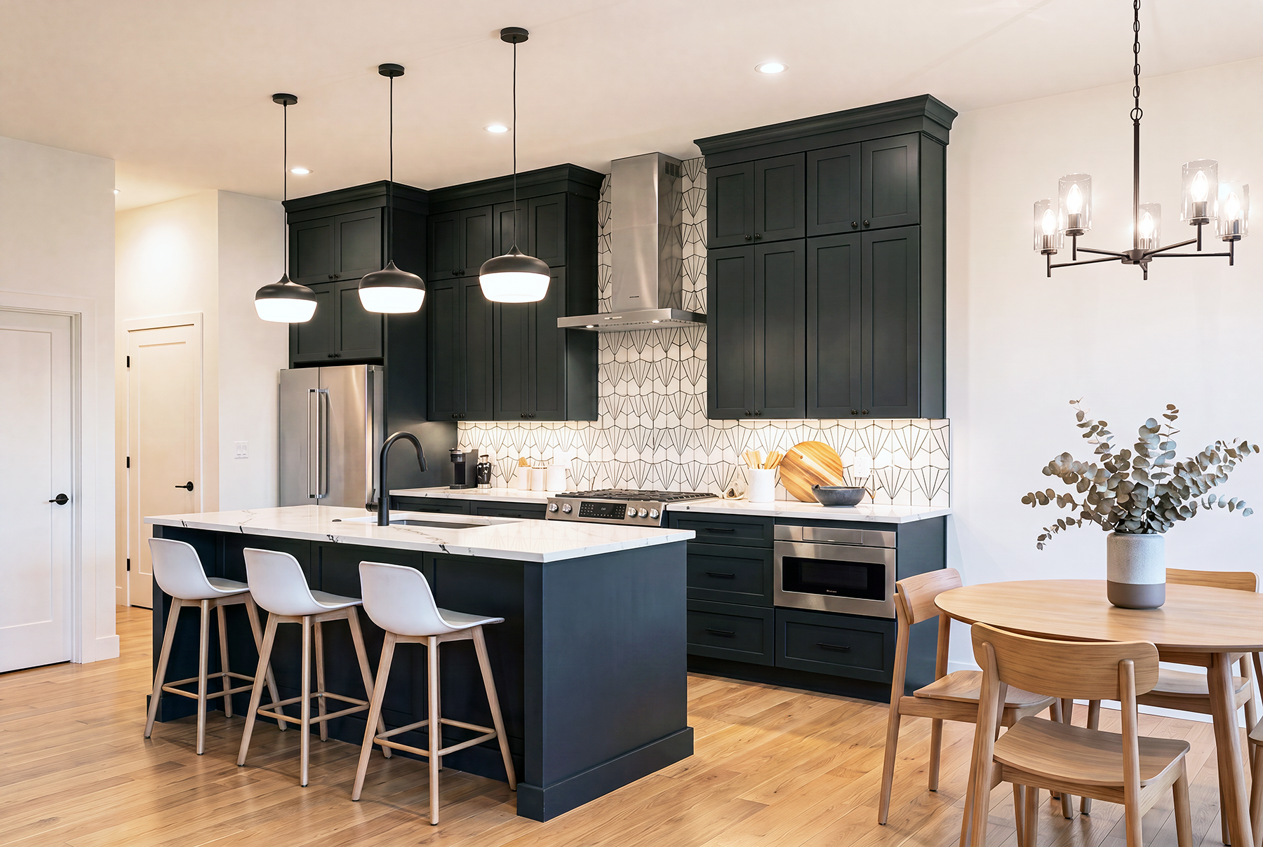 Modern kitchen with dark cabinets, a white marble island, three pendant lights, a stainless steel refrigerator, a geometric backsplash, and a round wooden dining table with chairs and a vase of eucalyptus