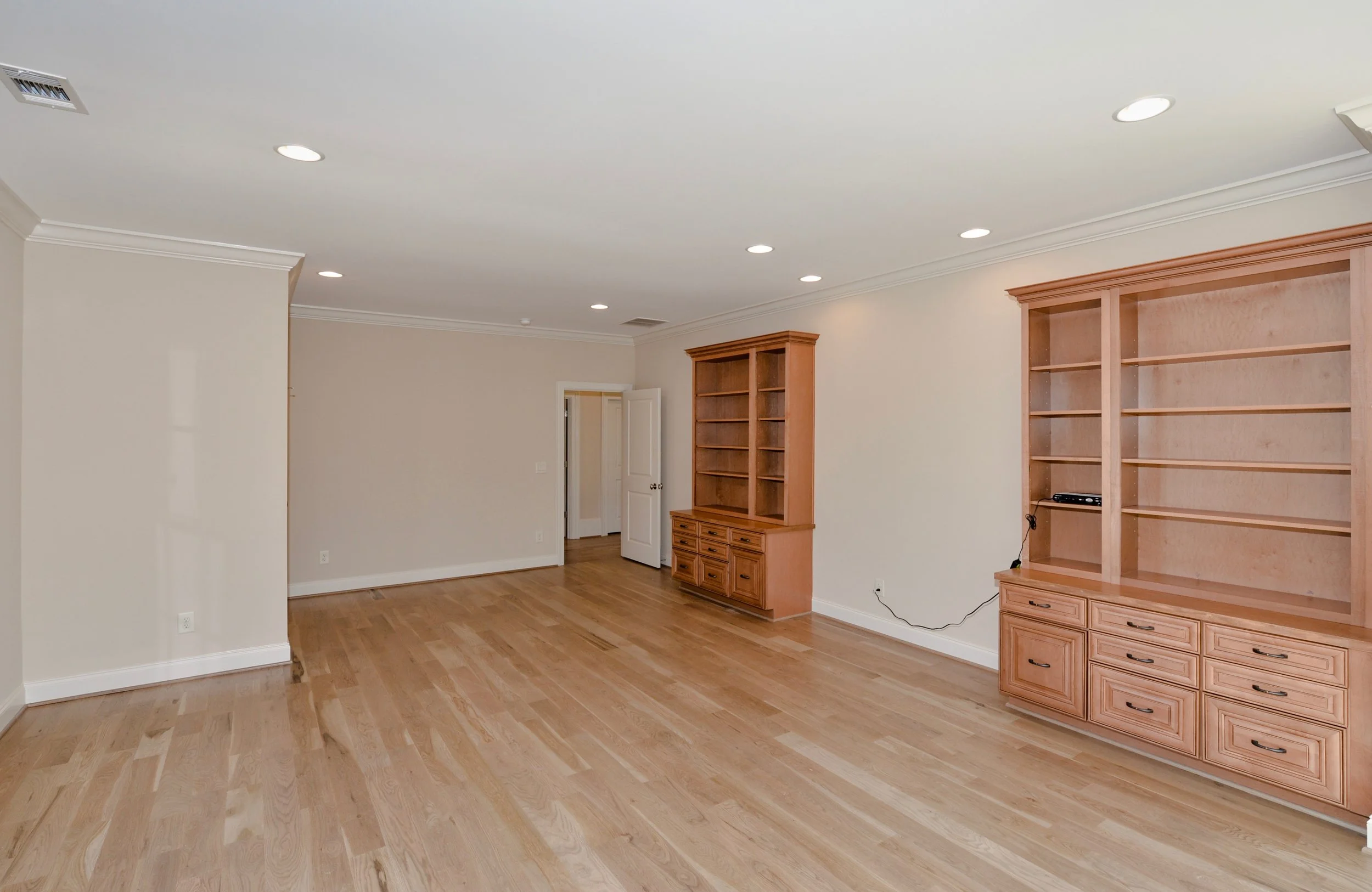 Empty living room with hardwood floors, beige walls, white trim, recessed lighting, and built-in wooden shelves and cabinets.