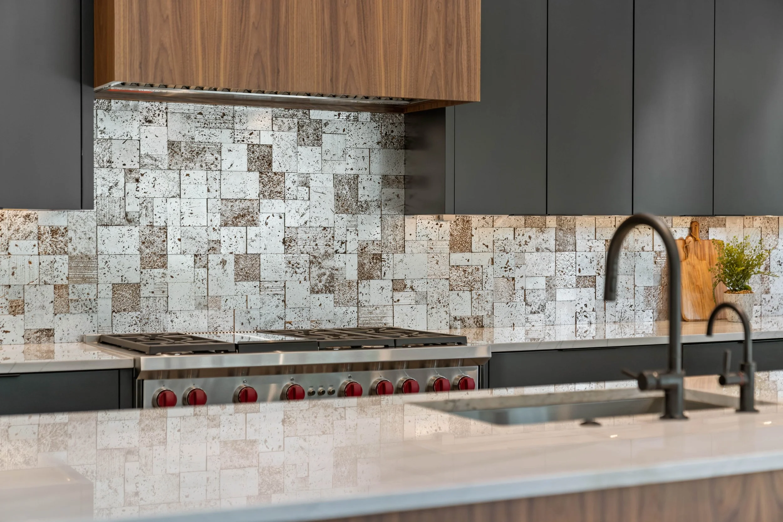 Modern kitchen with dark cabinetry, a mosaic tile backsplash in white and brown, a stainless steel stove with red knobs, and a white countertop with a black faucet and a small potted plant.