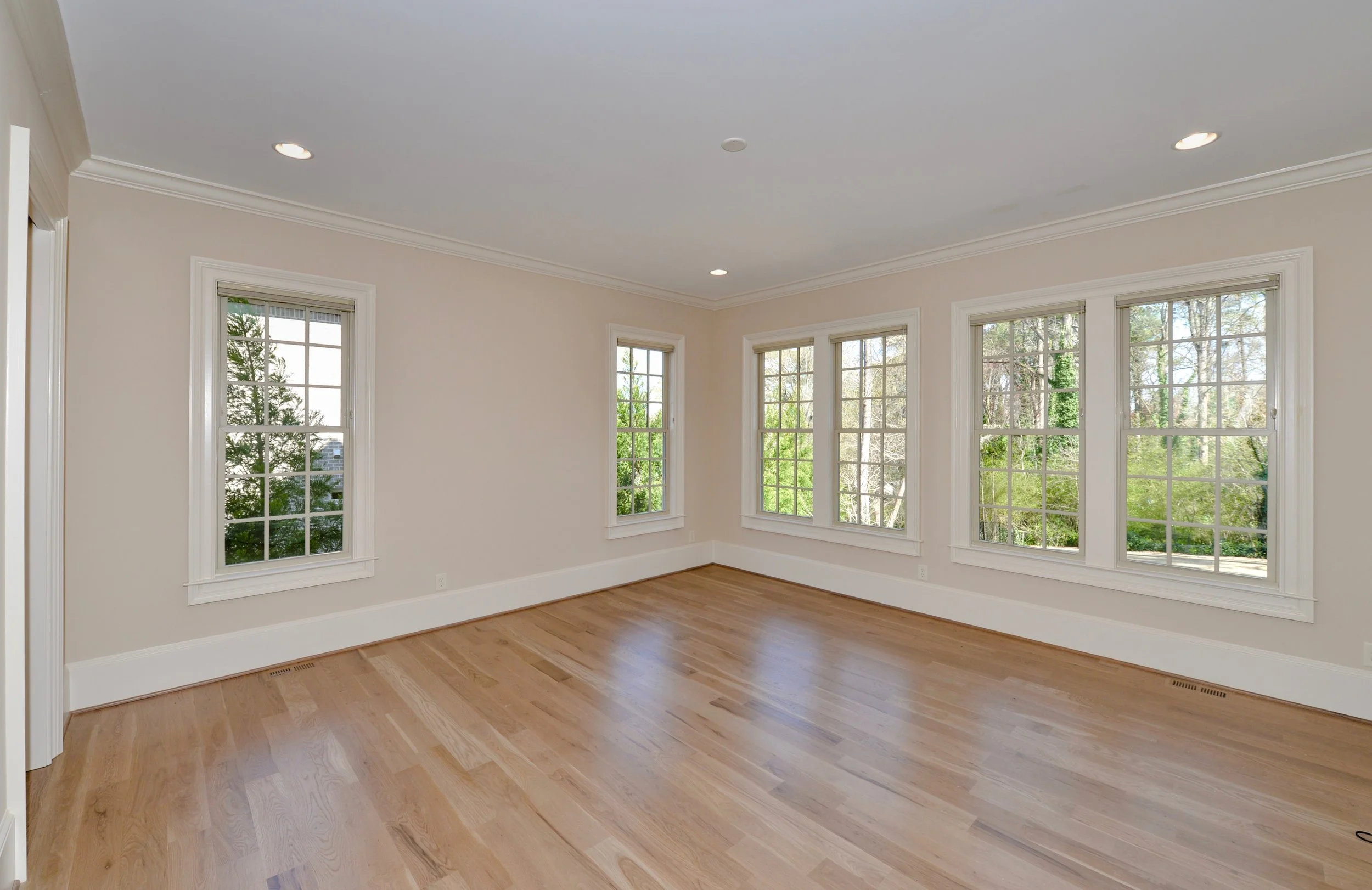 Empty living room with hardwood floors and multiple large windows allowing natural light, white walls, and crown molding.