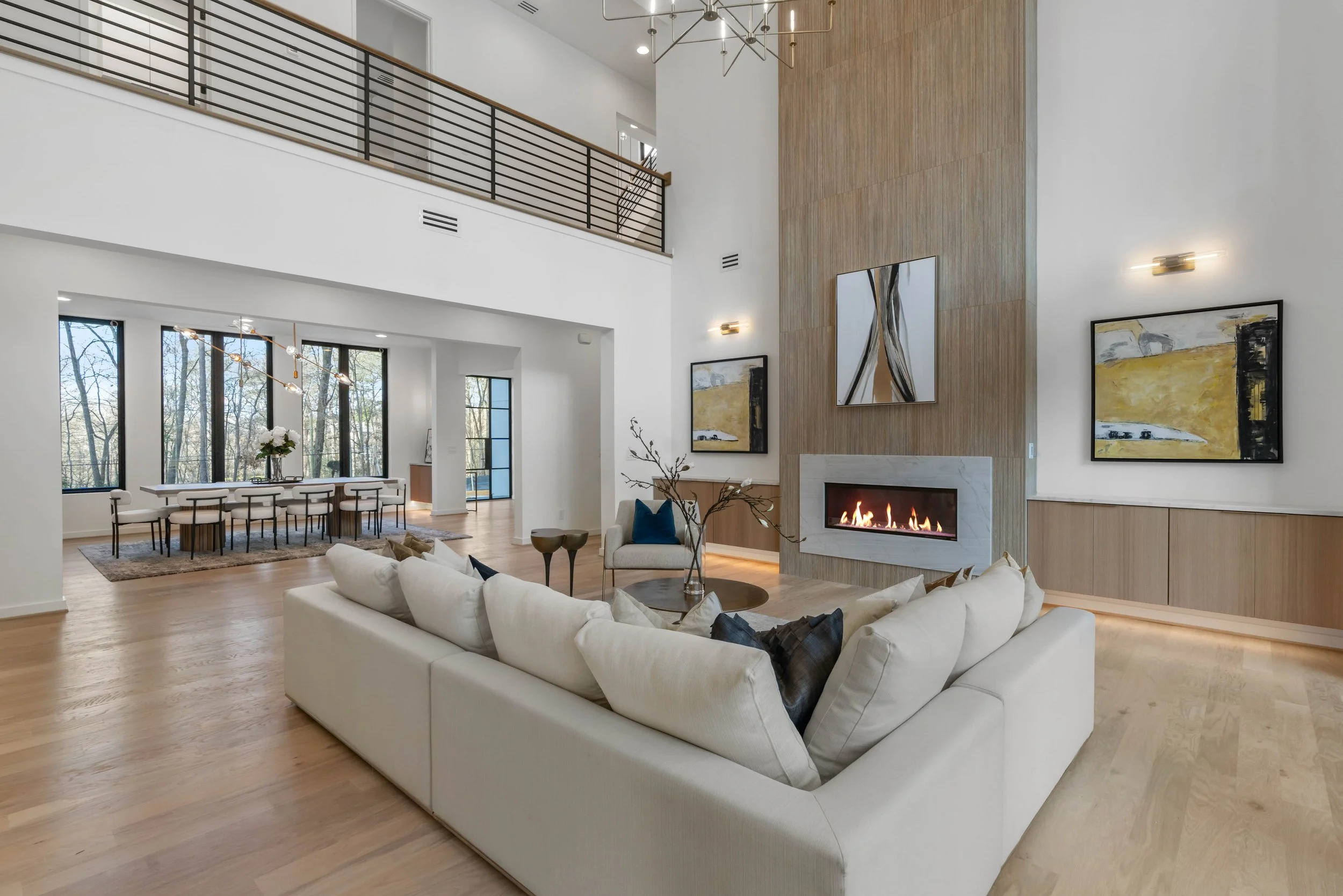 Modern living room with a white sectional sofa, abstract artwork above a fireplace, wooden accent wall, and a dining area with a long table and chairs in the background.