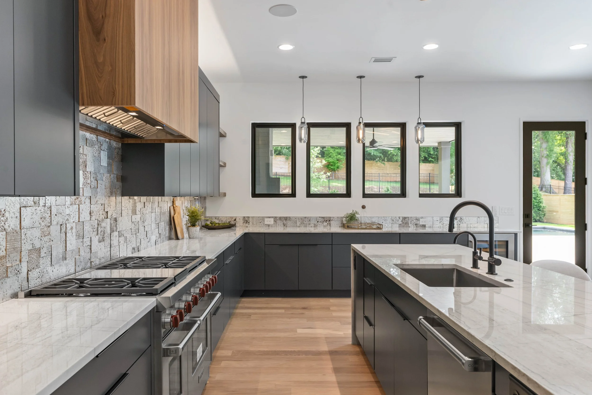 Modern kitchen with black cabinets, white marble countertops, a large island with a sink, four pendant lights, and large windows overlooking a backyard.