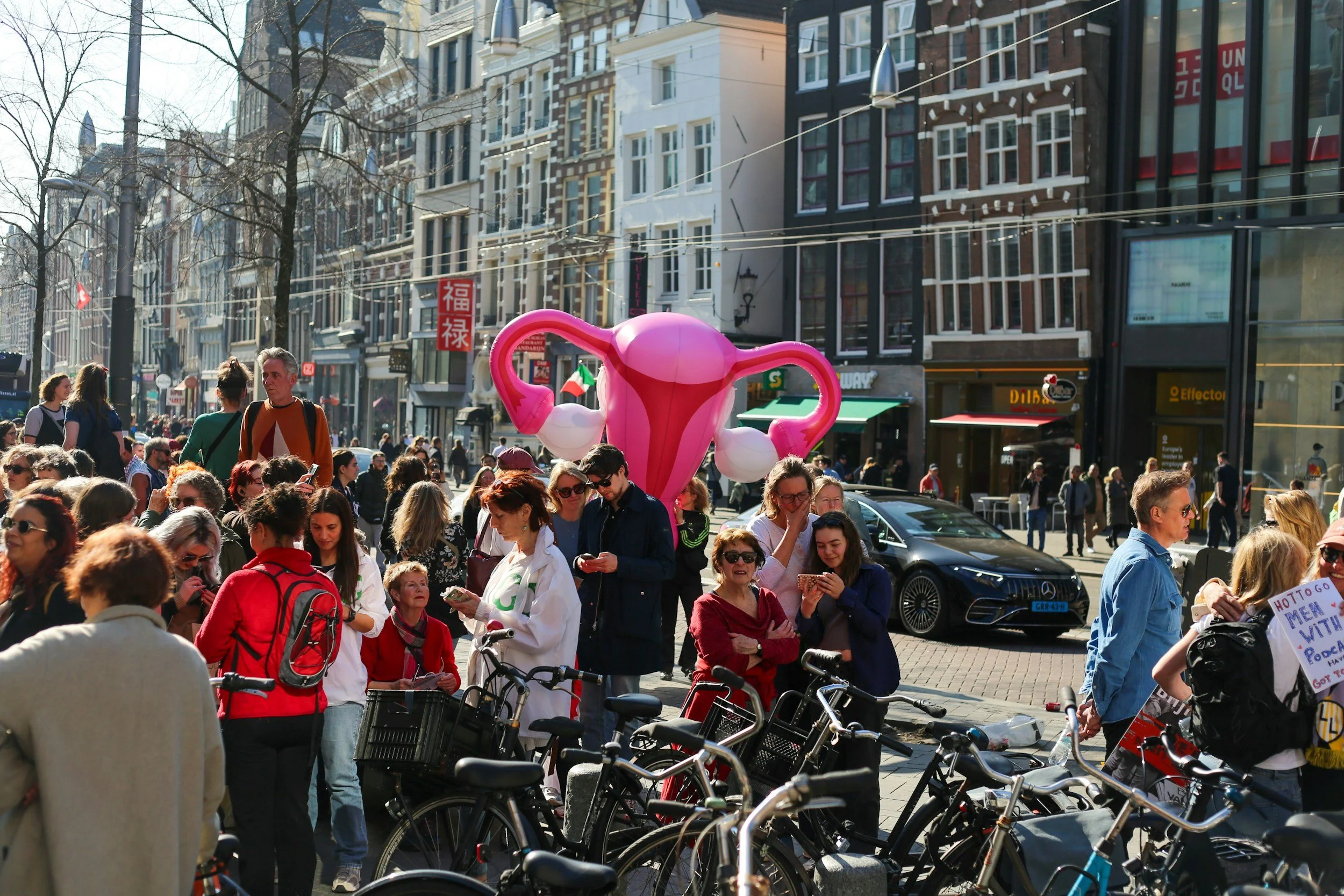 Crowded city street with people walking and biking, a large pink breast pump sculpture, and buildings in the background.