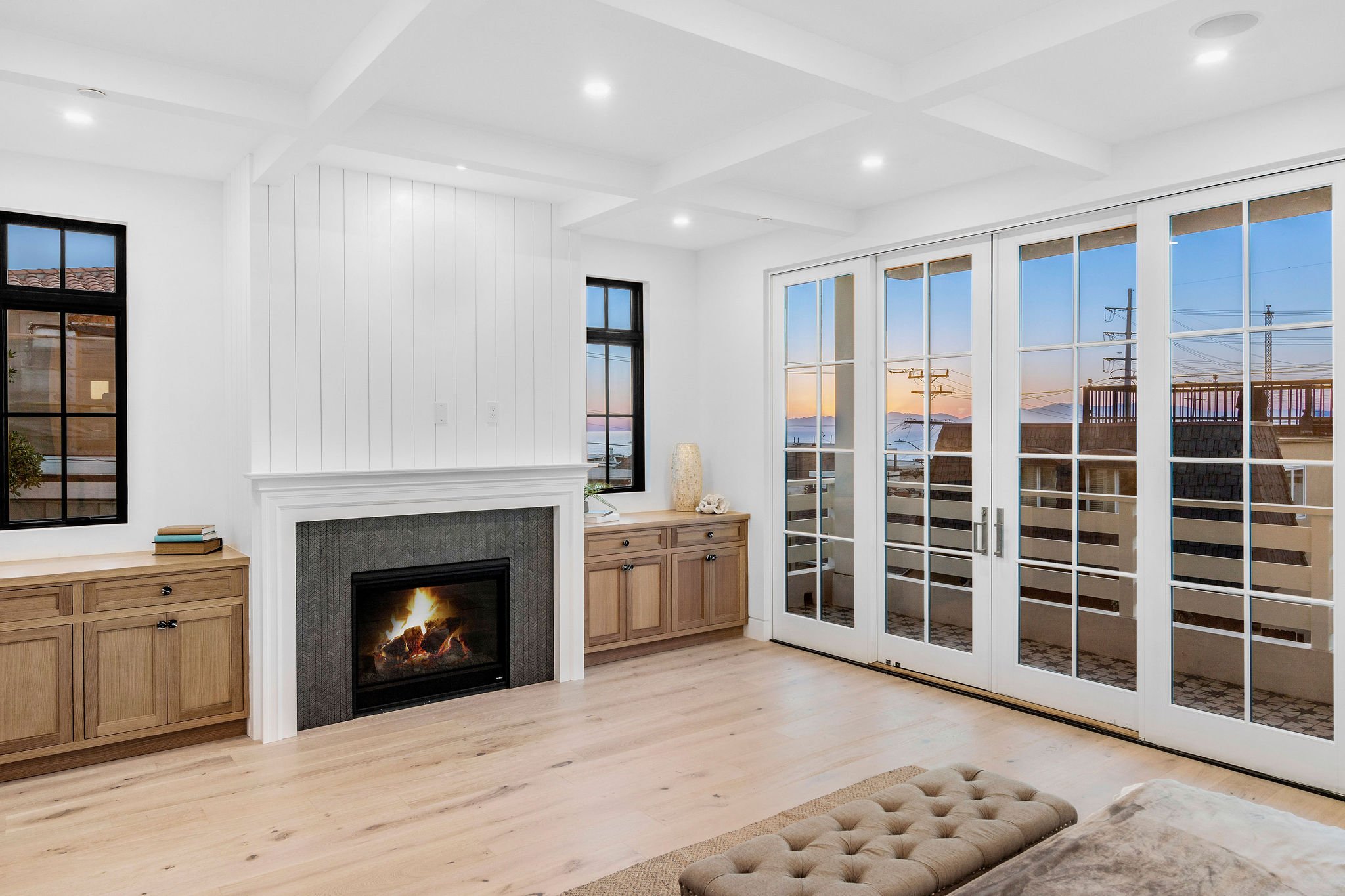 Modern living room with a fireplace, hardwood floors, and large glass doors leading to a balcony with a sunset view. Built-in cabinets and recessed lighting enhance the bright, cozy space.
