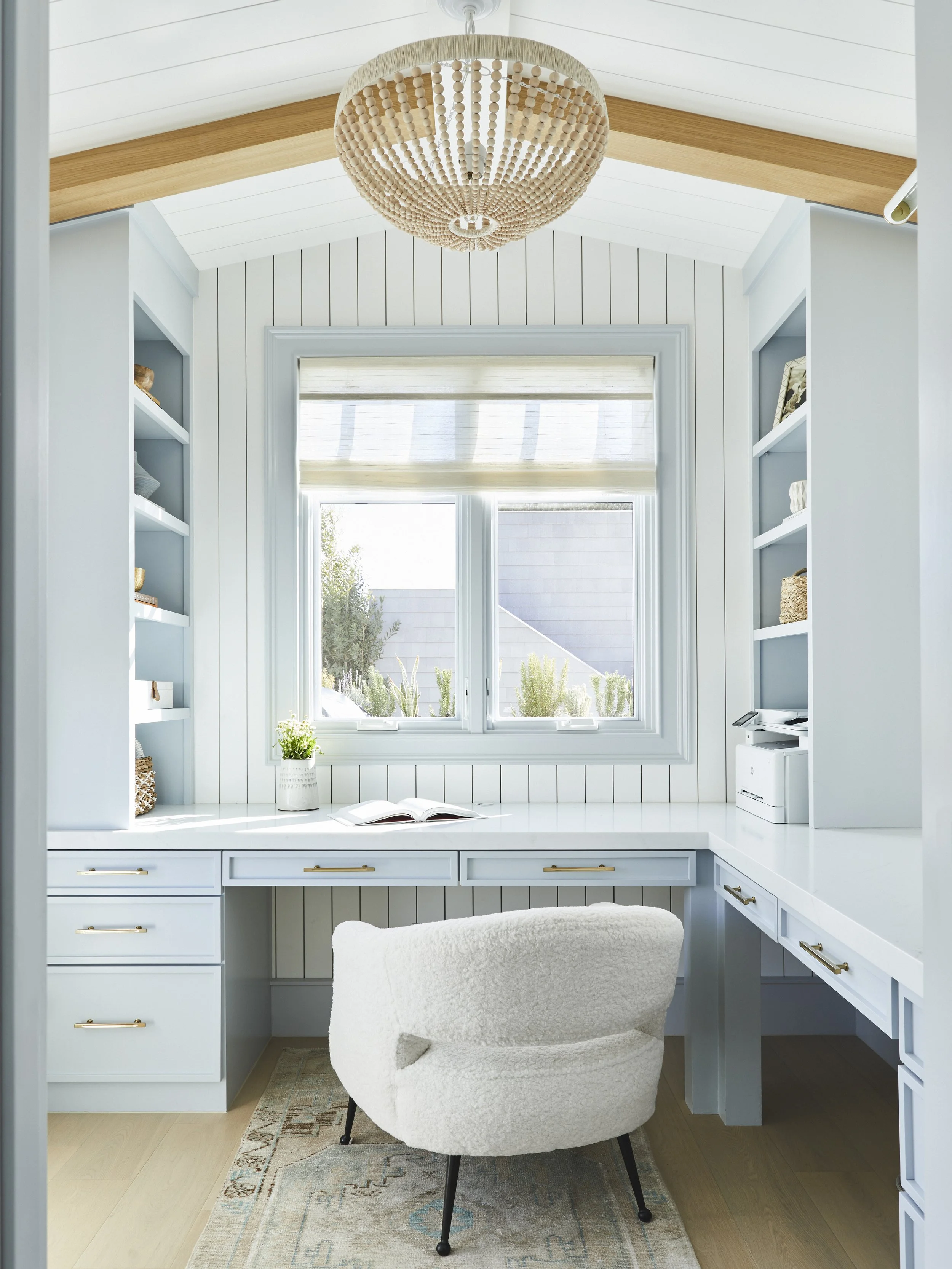 Bright home office with a white desk, open book, cozy chair, and large window. Light wood ceiling beams and a woven chandelier add warmth. Shelves hold decor items and a printer. Modern, airy design with neutral tones.