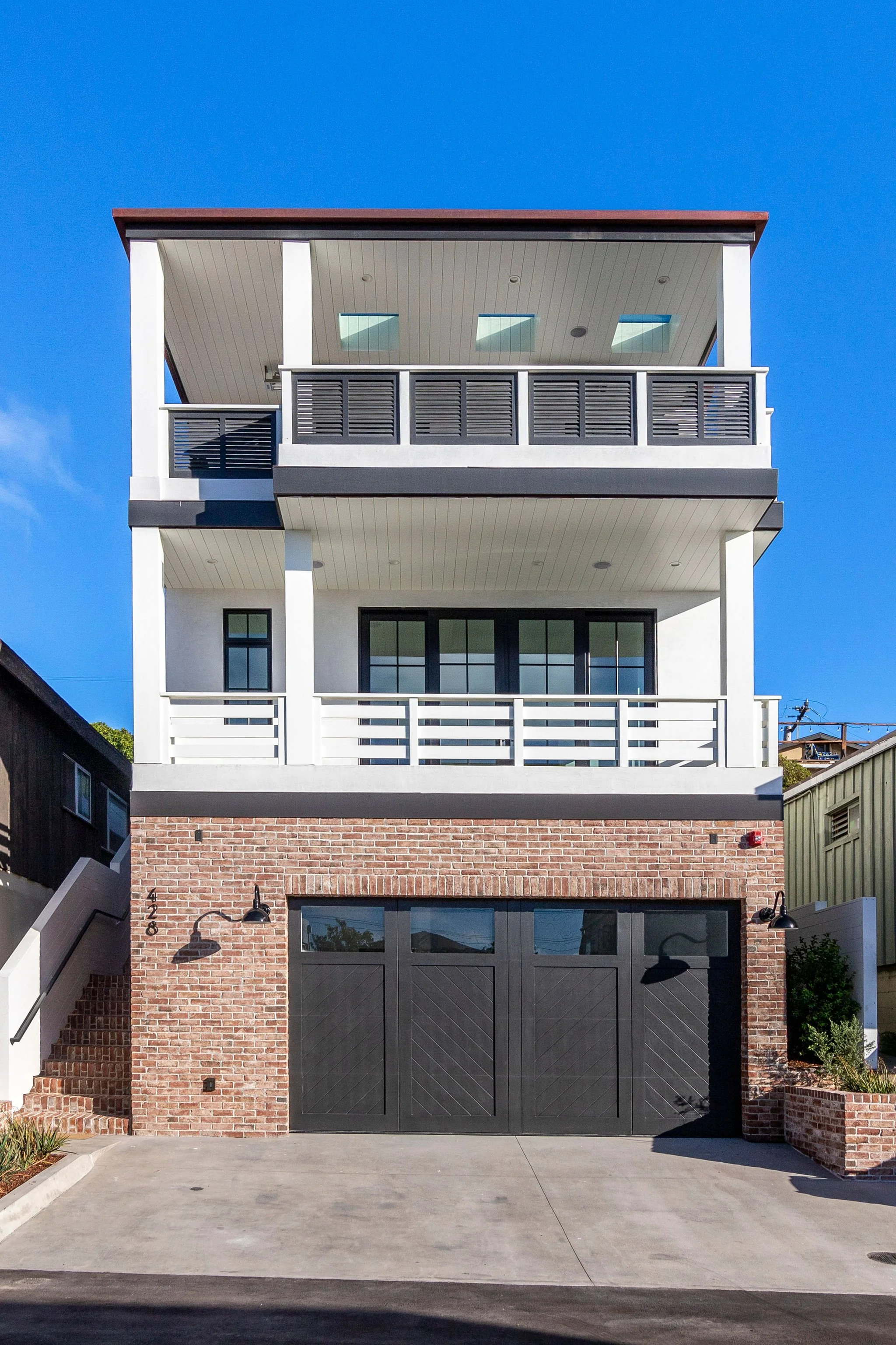 Modern three-story house with brick facade and black garage doors, featuring balconies and large windows under a clear blue sky.