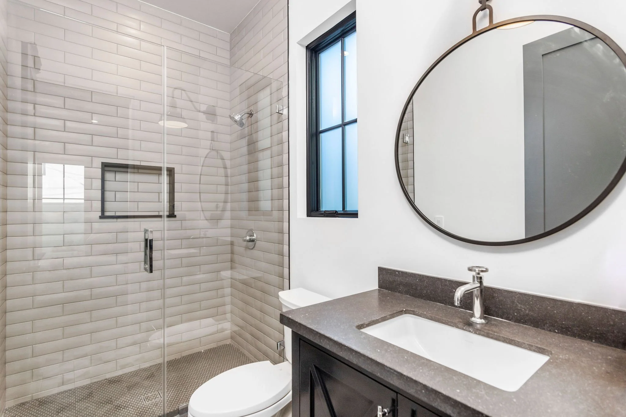 Modern bathroom featuring a glass-enclosed shower with subway tile, a round mirror, a dark countertop with a sink, and a toilet. There is a window with a black frame.