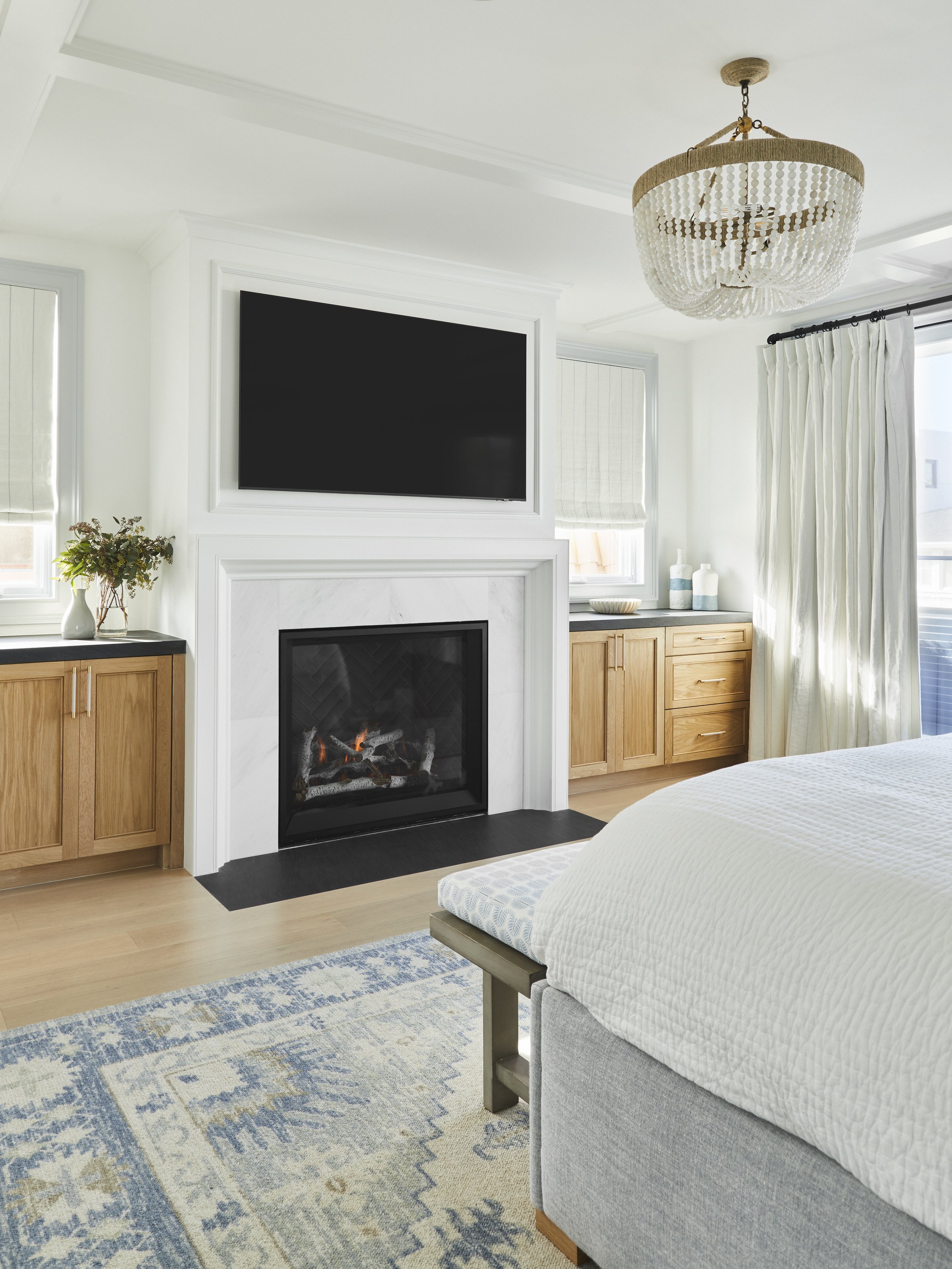A modern bedroom interior with a cozy fireplace, a wall-mounted TV, a decorative chandelier, wooden cabinetry, a large bed with a white comforter, and a patterned area rug. Two windows with Roman shades and light-colored curtains are present.