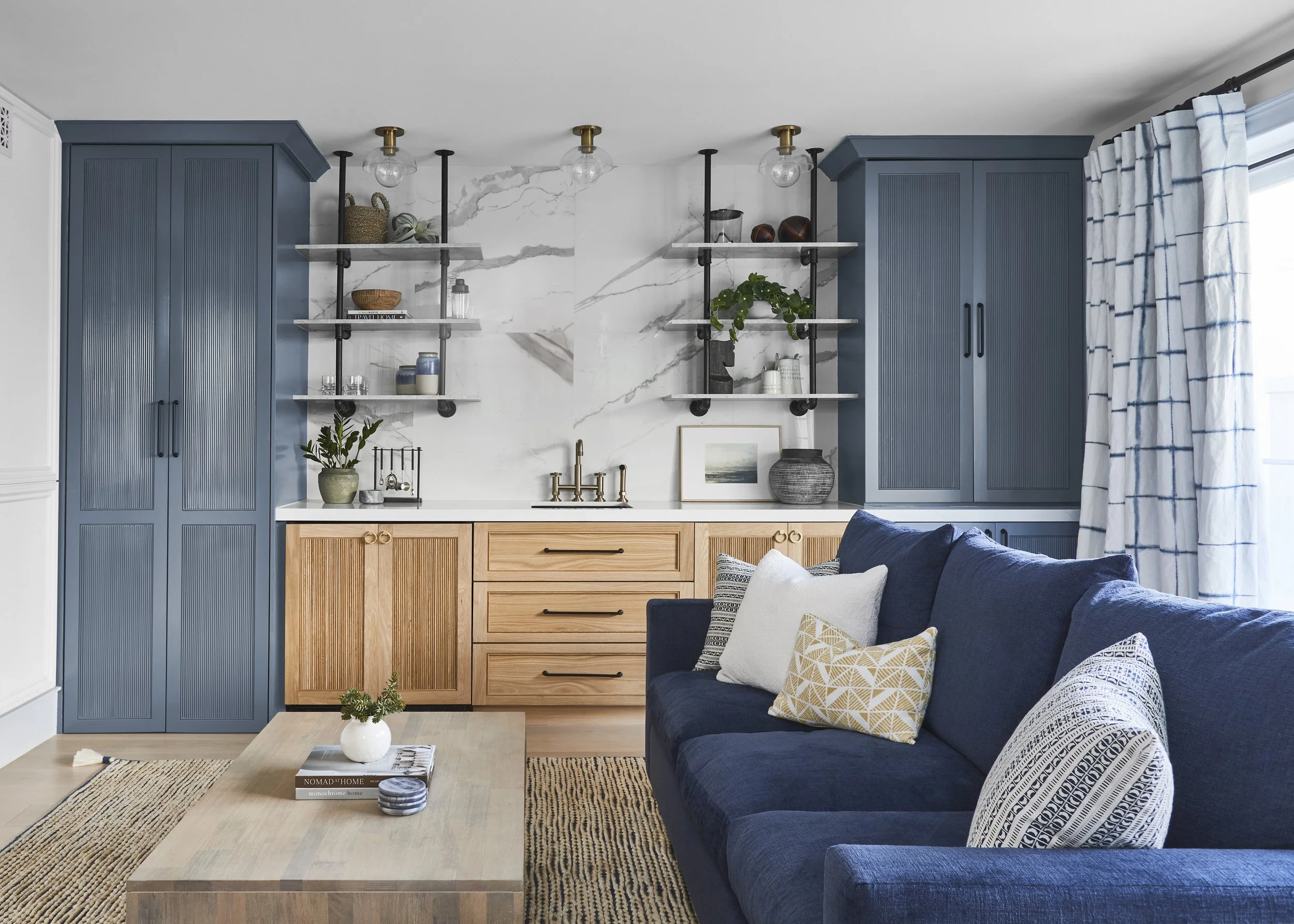 Stylish living room with a navy blue sofa, patterned pillows, light wood coffee table, and wood cabinets with blue accents. The wall features white marble patterns, open shelves with decor items, and a brass faucet. Light streams in through large win