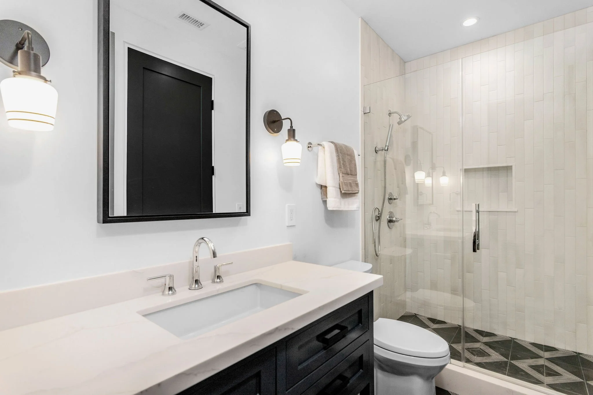 Modern bathroom with glass shower, black vanity, and white countertop, featuring a large mirror, wall light fixtures, and a towel rack with towels.