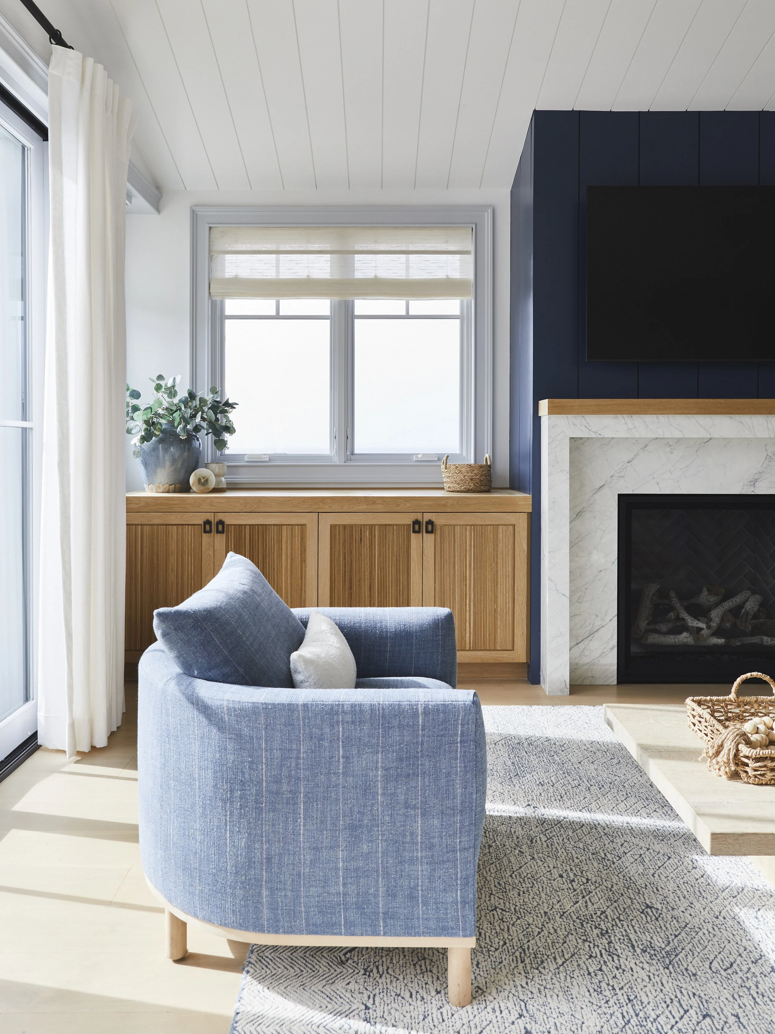 Modern living room with blue armchair, beige rug, wooden cabinets, indoor plant, fireplace, TV above mantel, and window with sheer curtains.