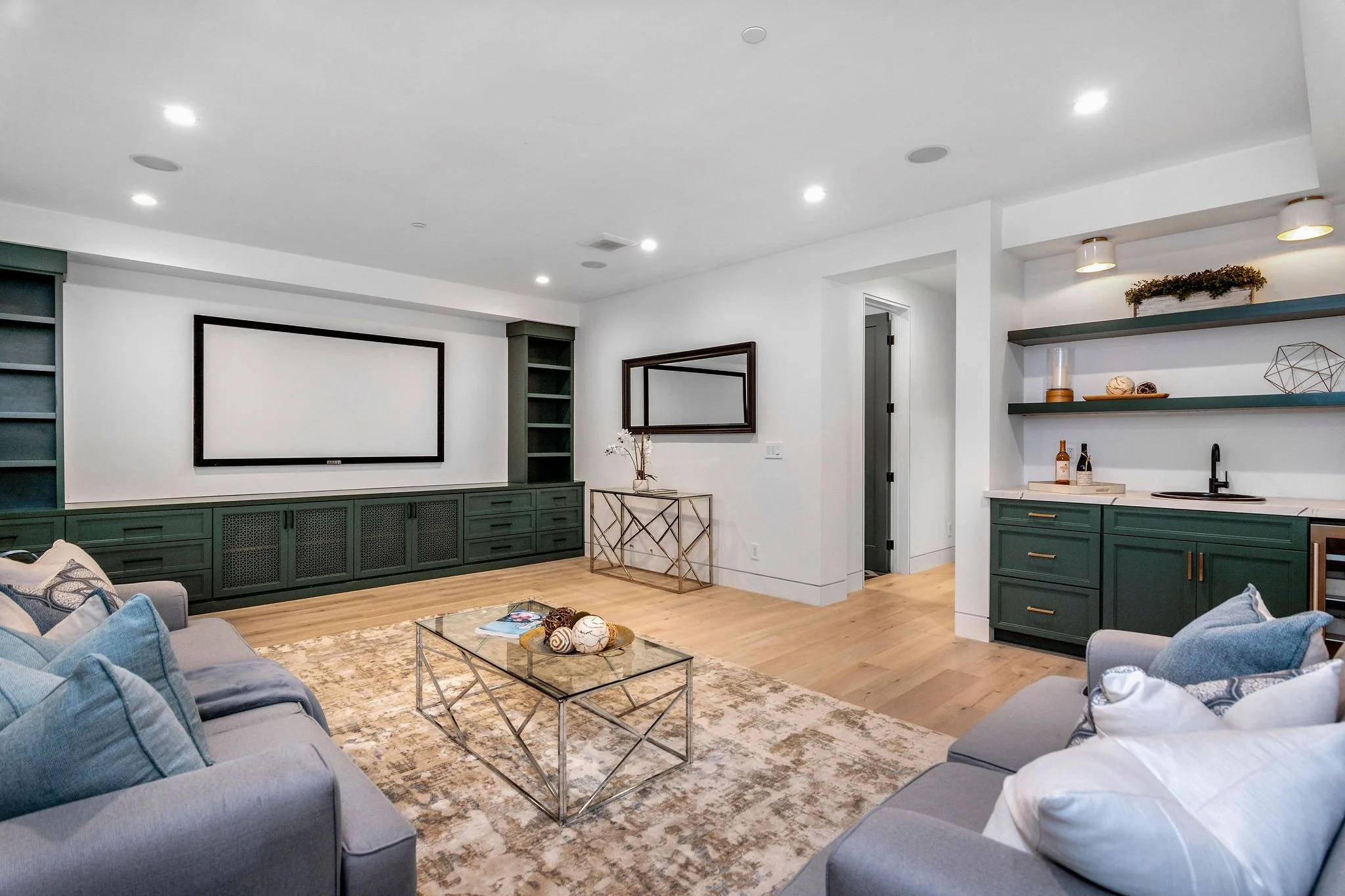 Modern living room with green cabinetry, large wall-mounted TV, two gray sofas, glass coffee table, light rug, wet bar with shelves, and decorative accents.