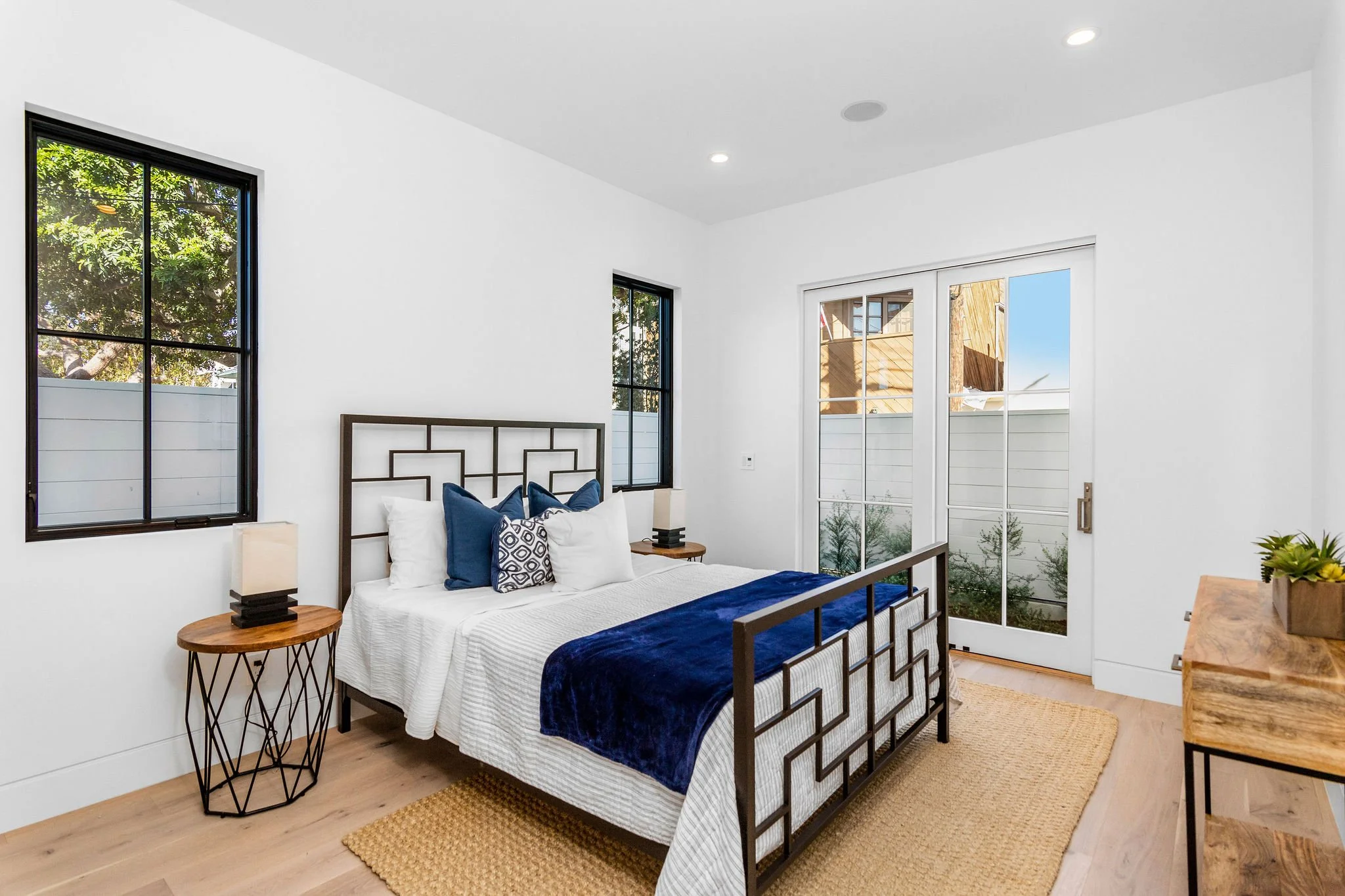 Modern bedroom with white walls, dark metal bedframe, blue and white bedding, wooden side tables, and natural light from large windows.