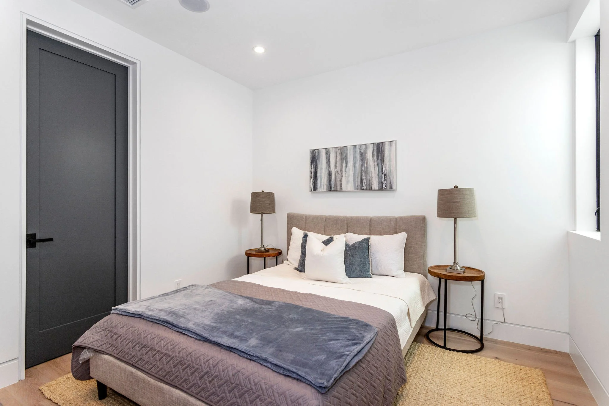 Modern bedroom with a gray door, a bed with a tufted headboard, white and gray bedding, two side tables with lamps, and abstract wall art.