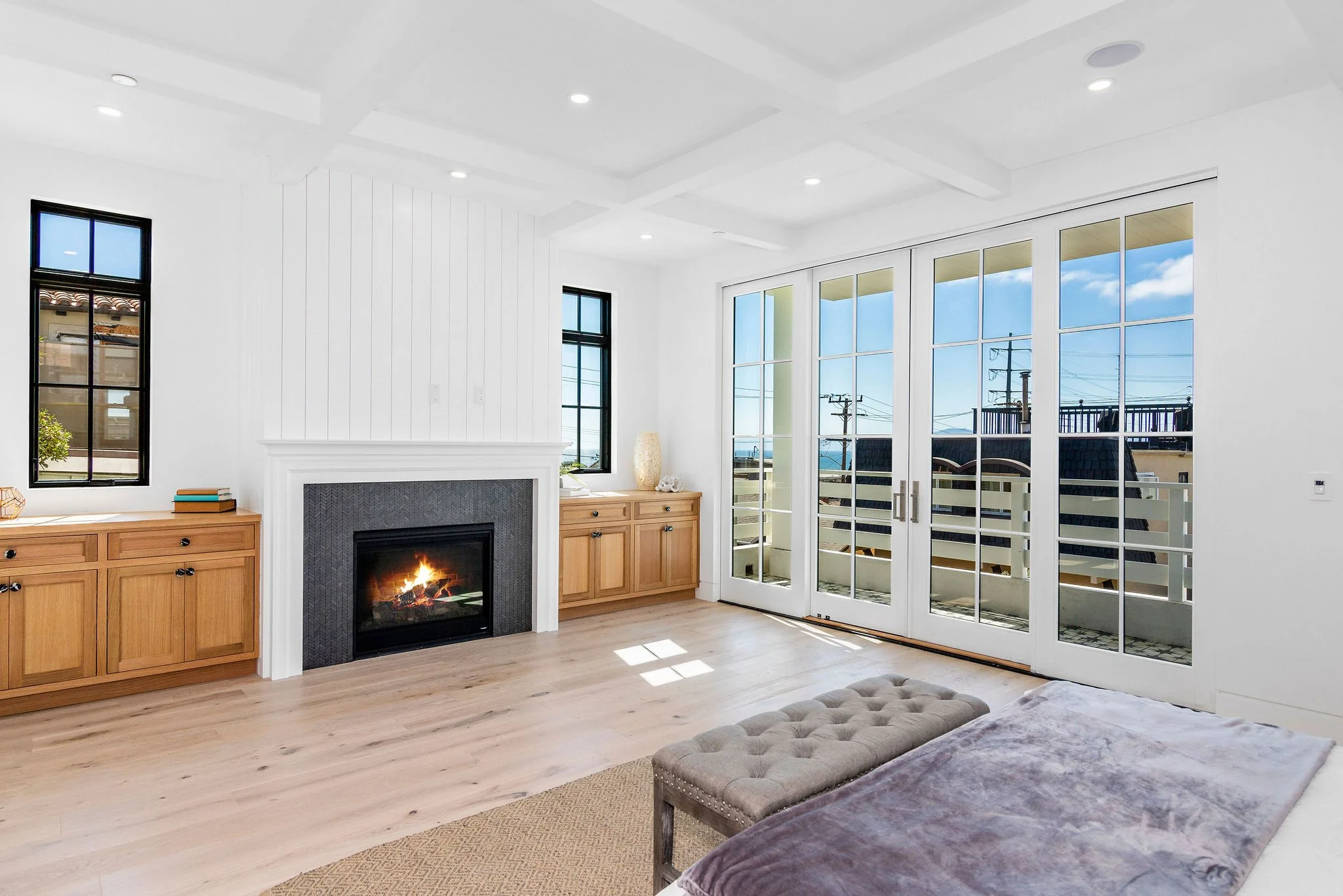 Bright living room with hardwood floors, a modern fireplace, large windows, glass double doors leading to a balcony, wooden cabinetry, and a tufted bench.