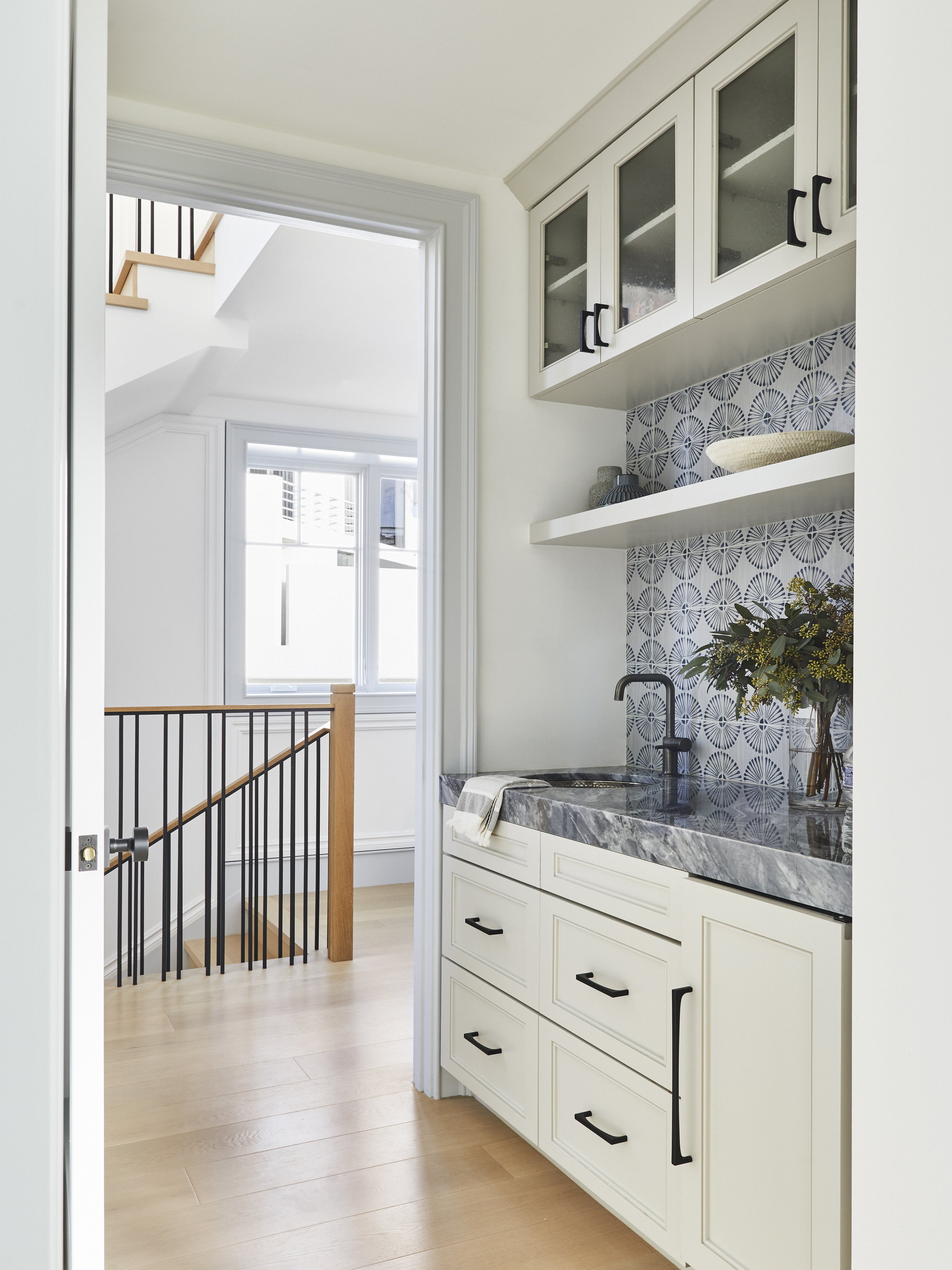 Small kitchen with patterned backsplash, white cabinets, marble countertop, and a sink with a black faucet. Wooden staircase in the background.