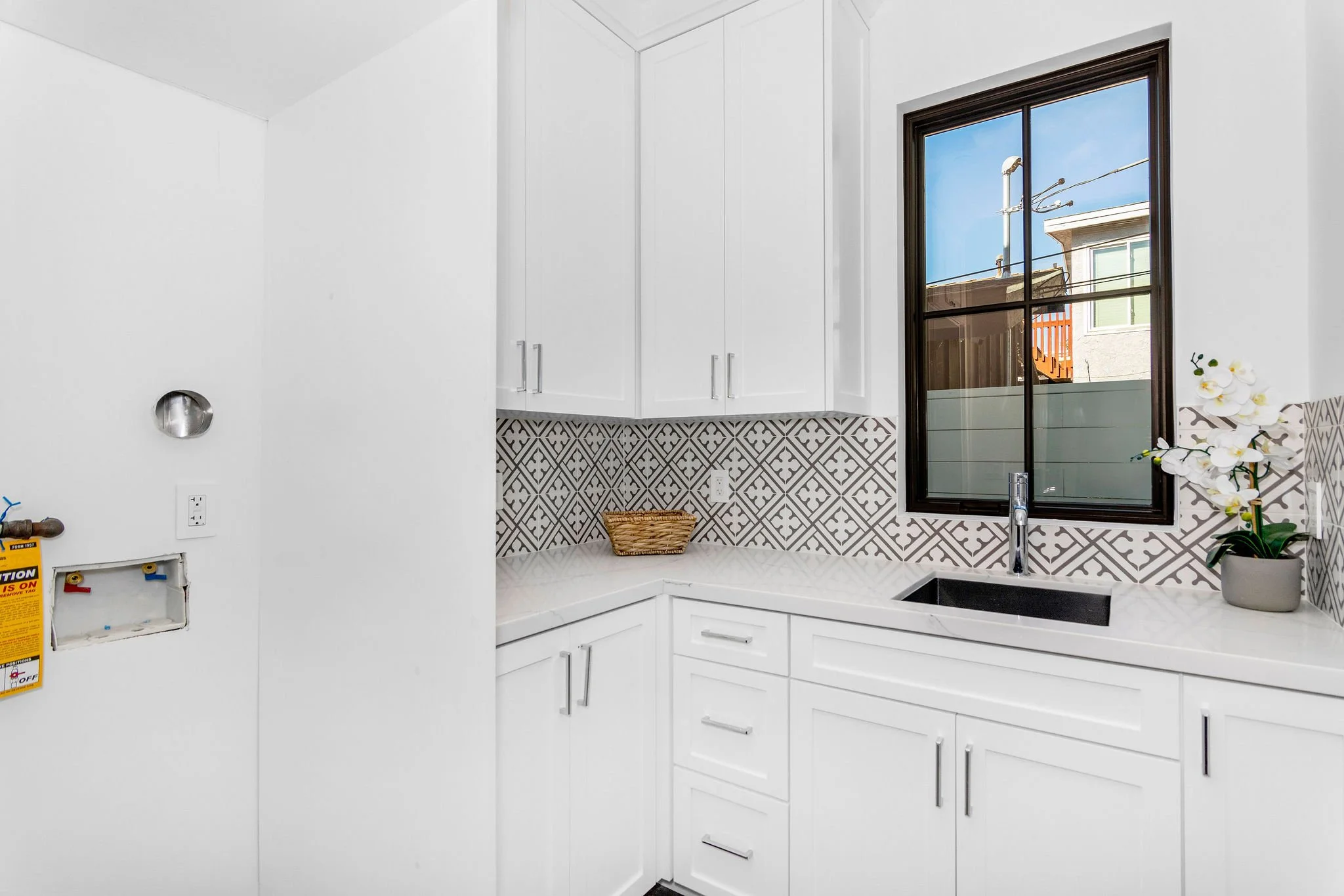 Laundry room with white cabinets, patterned tile backsplash, wall-mounted faucet, and a window.