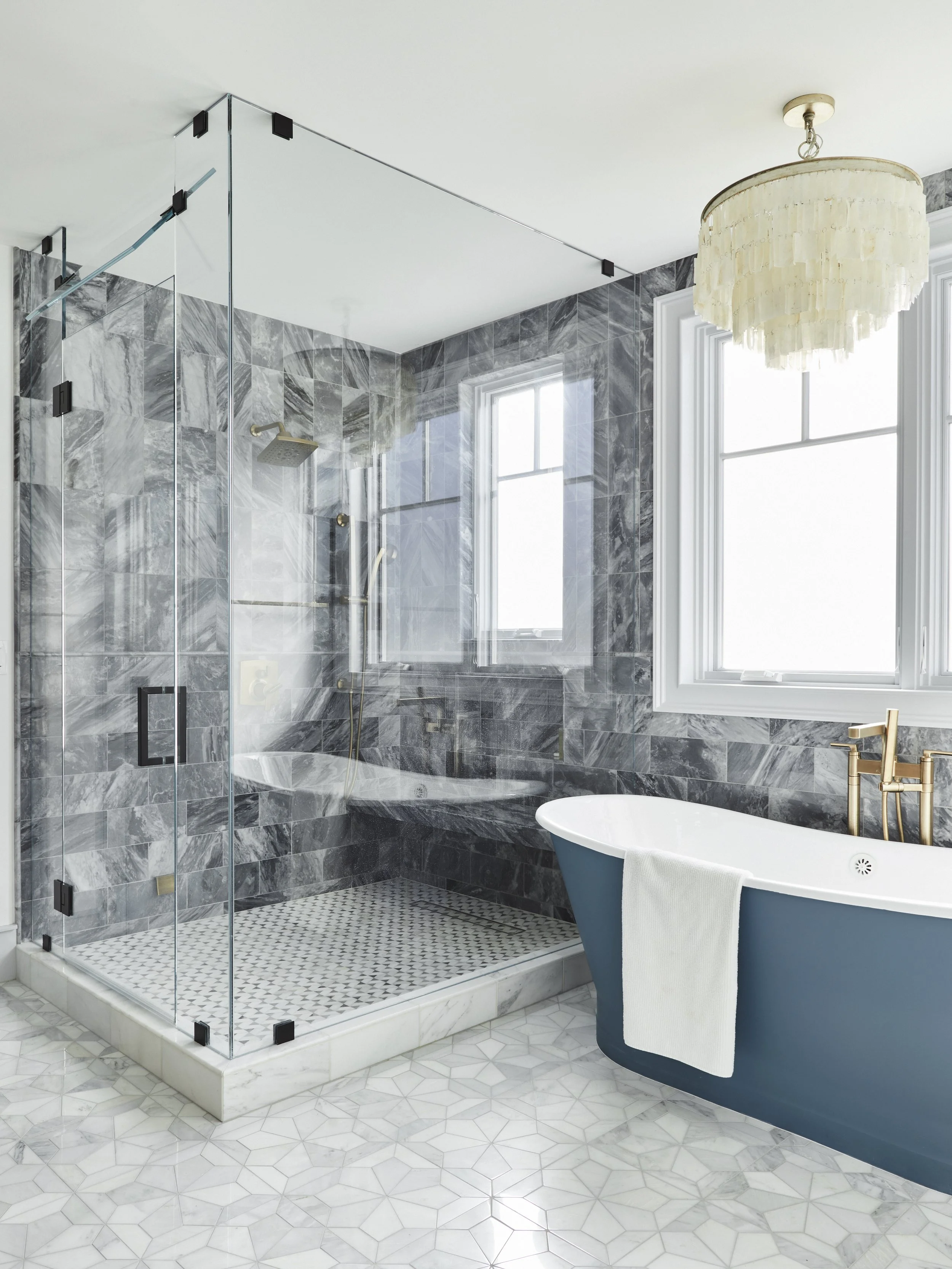 Modern bathroom with glass shower enclosure, freestanding bathtub, marble tile, and elegant chandelier.