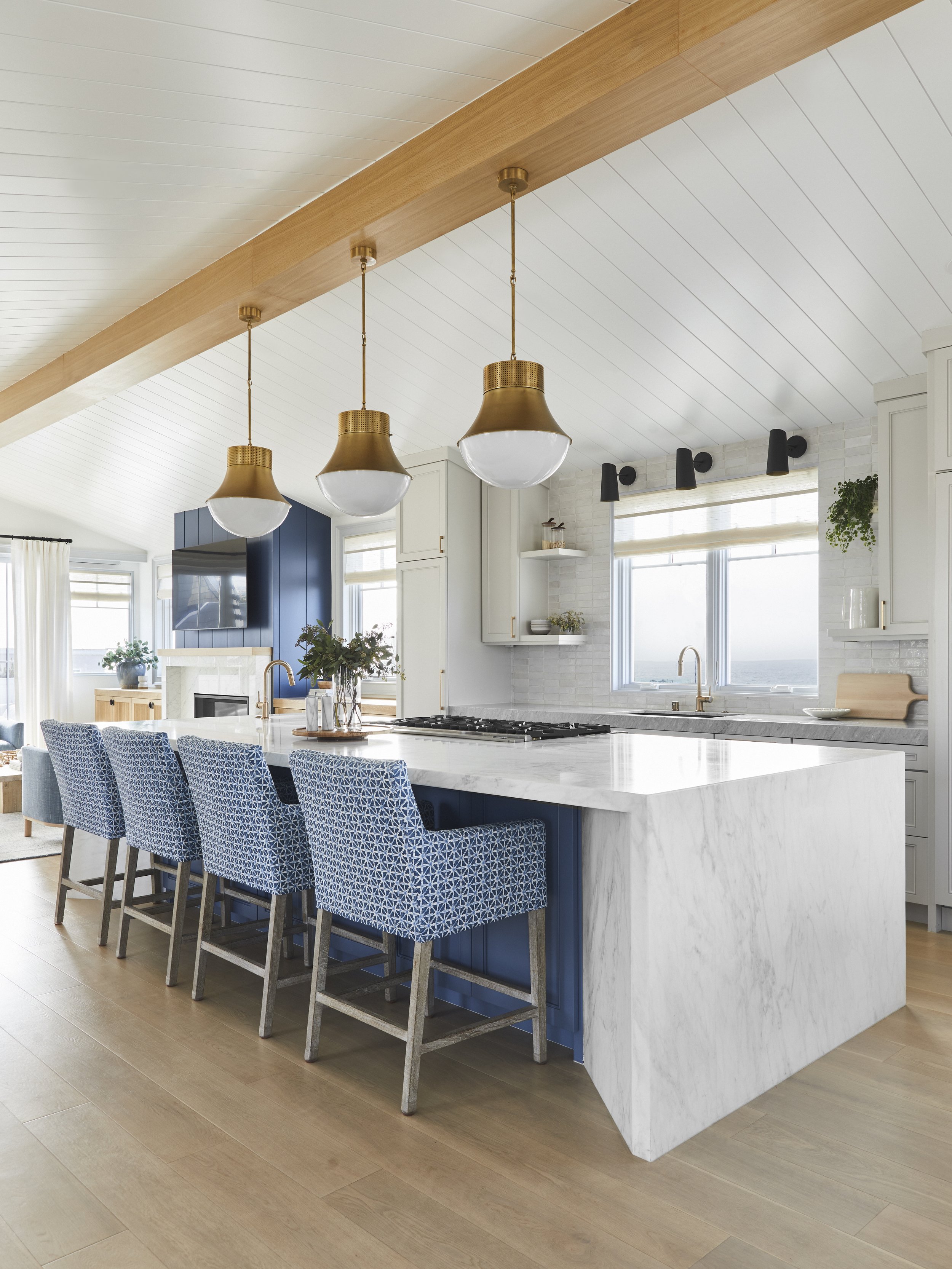 Modern kitchen interior with a large marble island, blue patterned bar stools, and brass pendant lights. The room features light wood flooring, white paneled ceiling, and a window with a view. Cabinets are white, and there are decorative plants on th