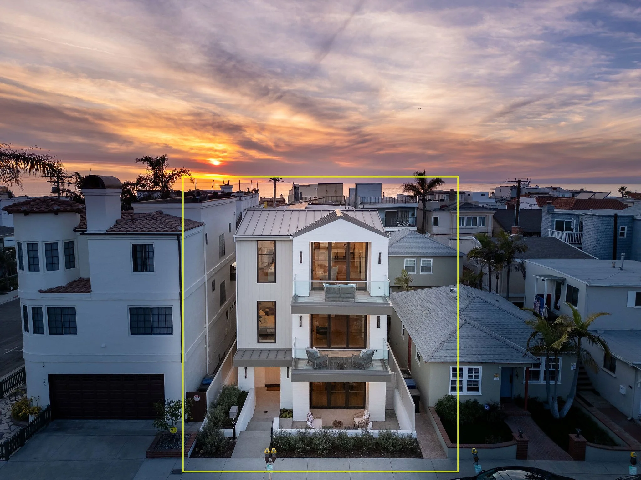 Modern multi-story house with glass windows, surrounded by other residential buildings, and palm trees at sunset.