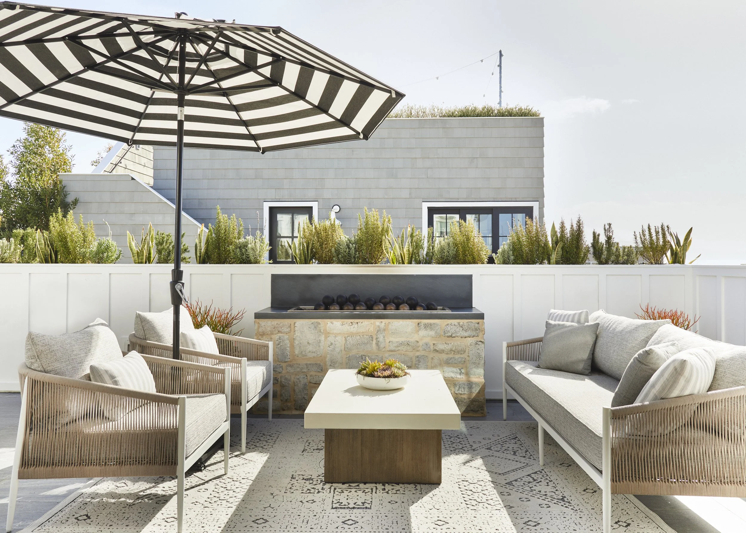 Outdoor patio with striped umbrella, wicker chairs, stone fireplace, and wooden table on a patterned rug.