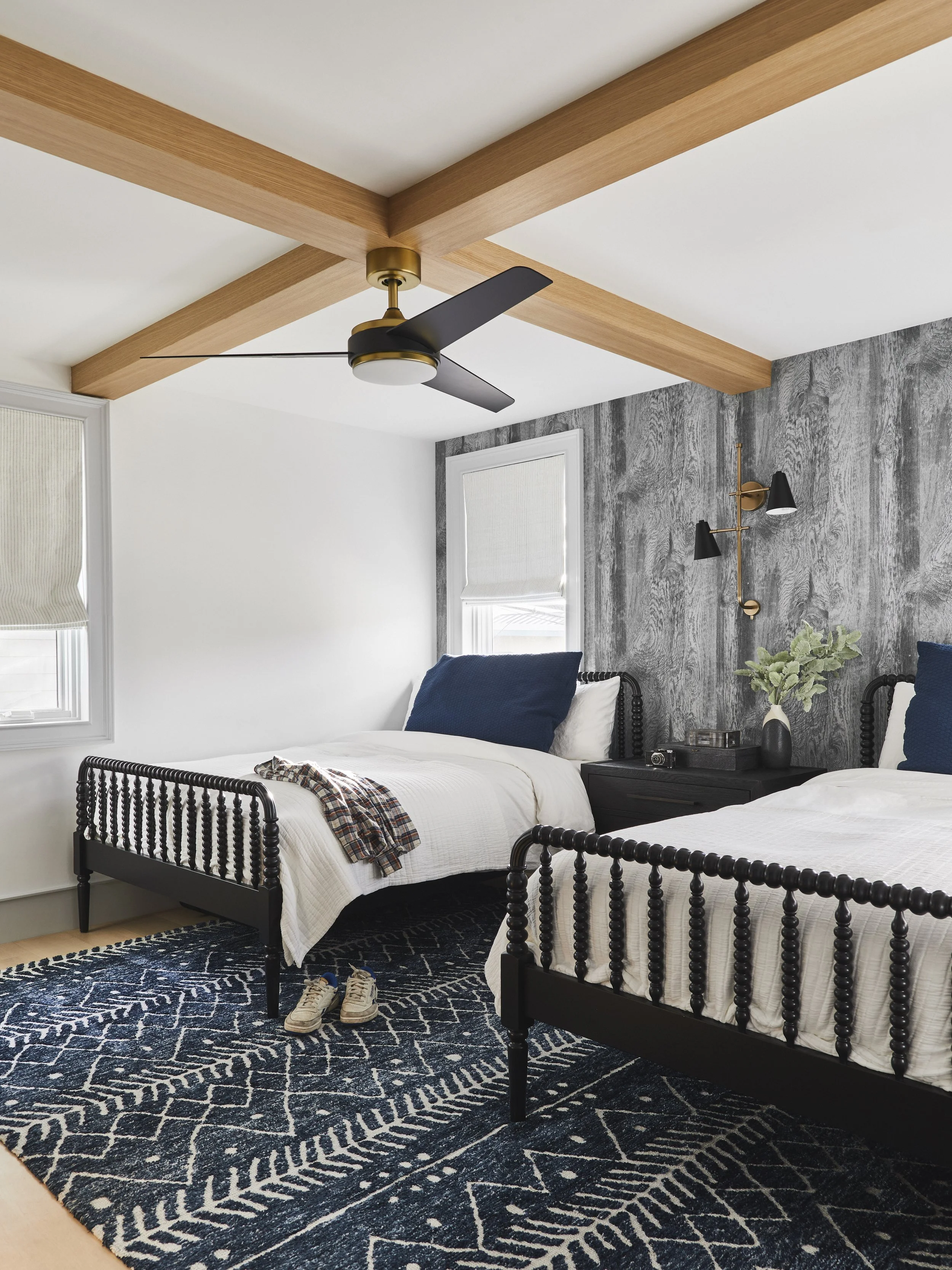 Bedroom with two twin beds featuring black spindle-style frames, a ceiling fan, and a blue patterned rug. The walls have a textured gray finish, and a plaid shirt is draped over one bed. A pair of sneakers rests on the rug. Light and dark blue pillow
