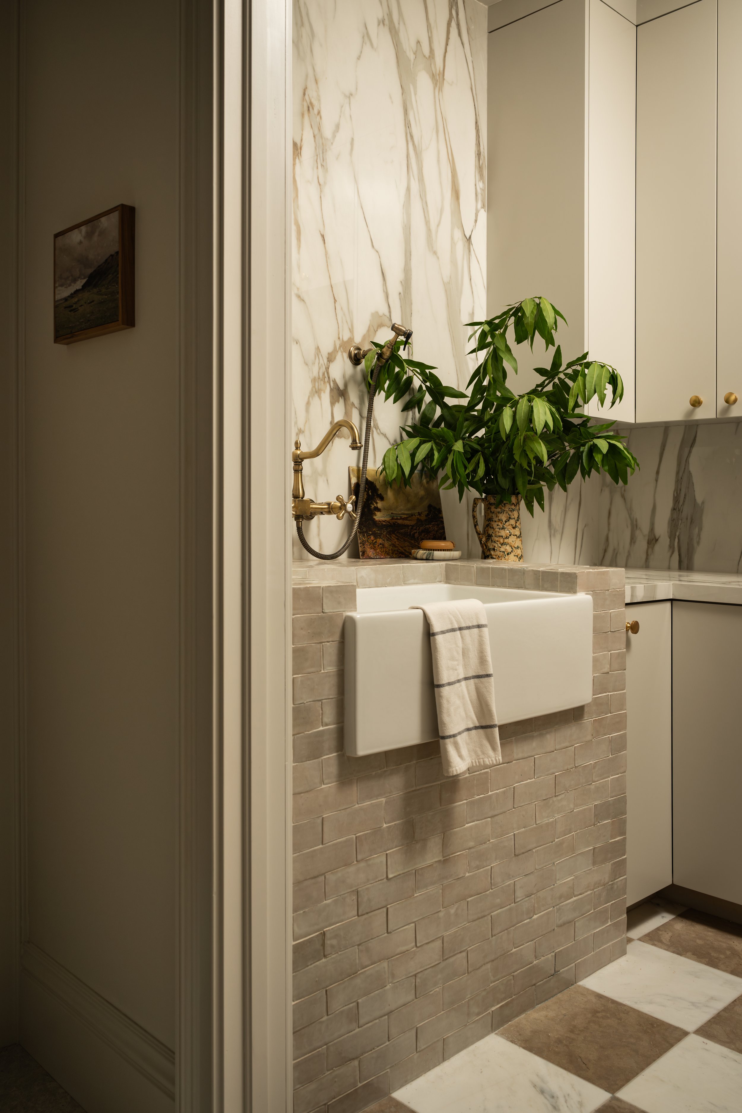 Modern bathroom with a marble wall, white sink, and brass faucet; potted plant on the counter, tiled walls, and a checkered floor. A towel hangs over the sink edge.