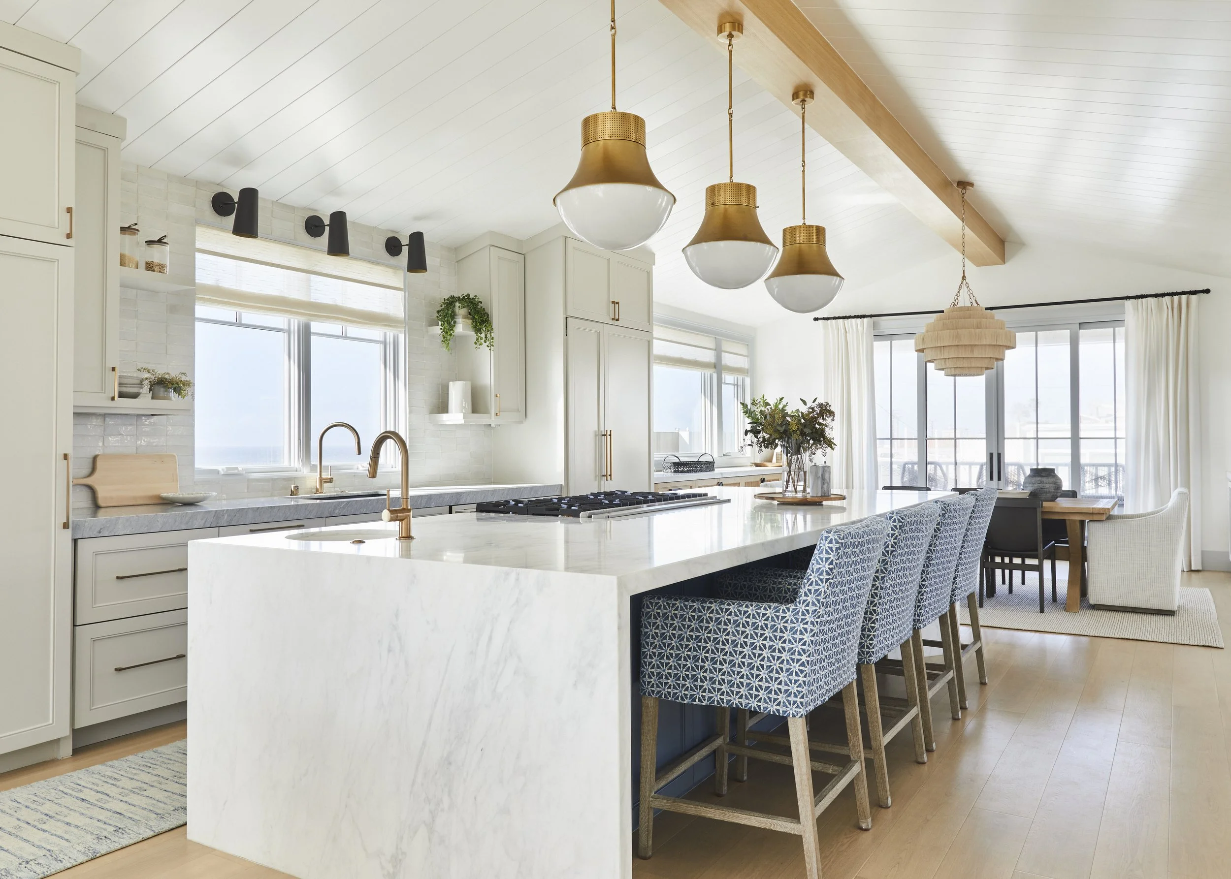 Modern kitchen with marble island, blue patterned chairs, wooden ceiling beam, pendant lights, large windows, and dining area.