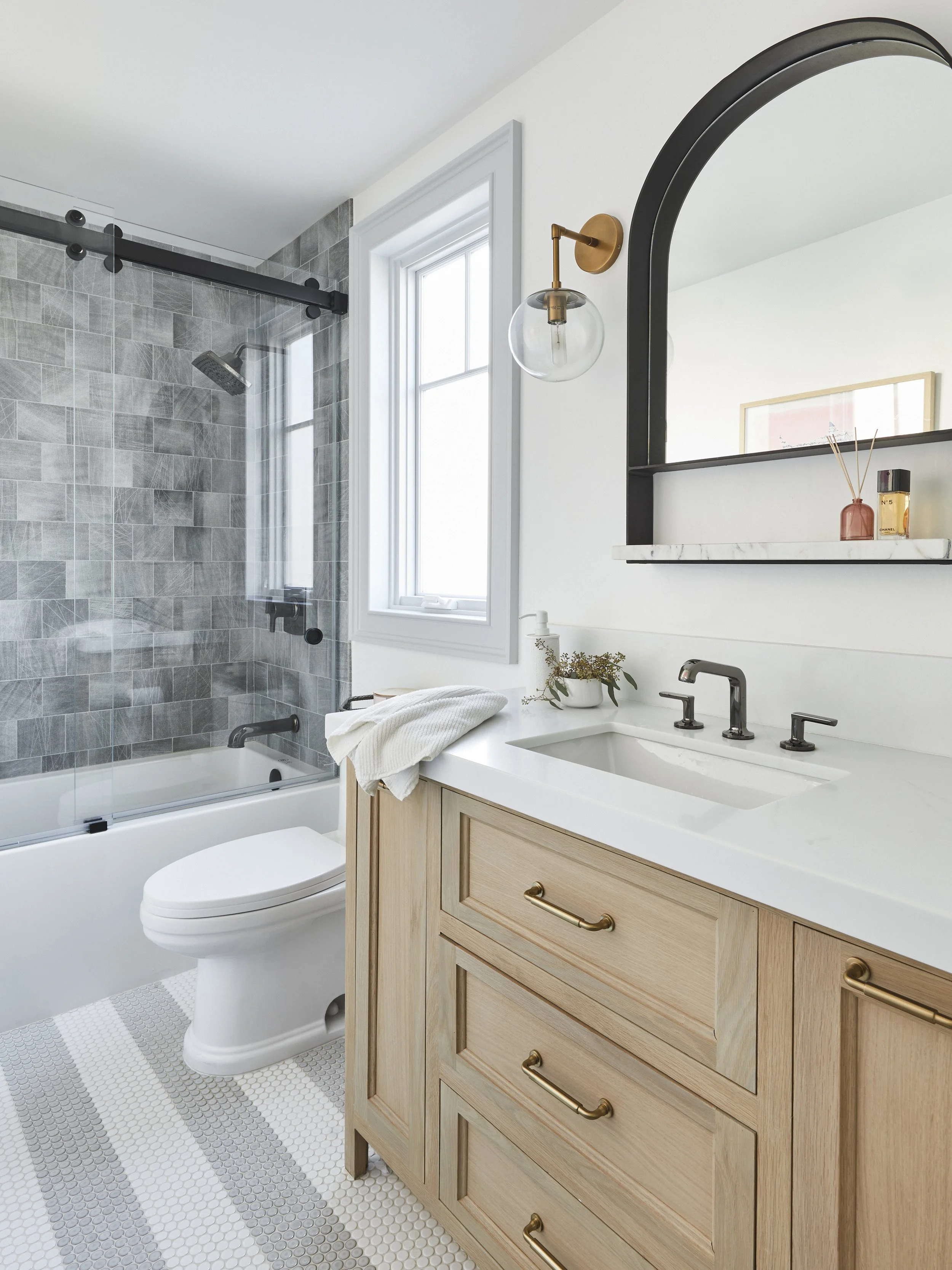 Modern bathroom with light wood vanity, white countertop, and brass fixtures, featuring a round wall sconce and an arch mirror. The bathroom includes a glass-enclosed bathtub with gray tile, a large window, and a striped hexagonal tile floor.