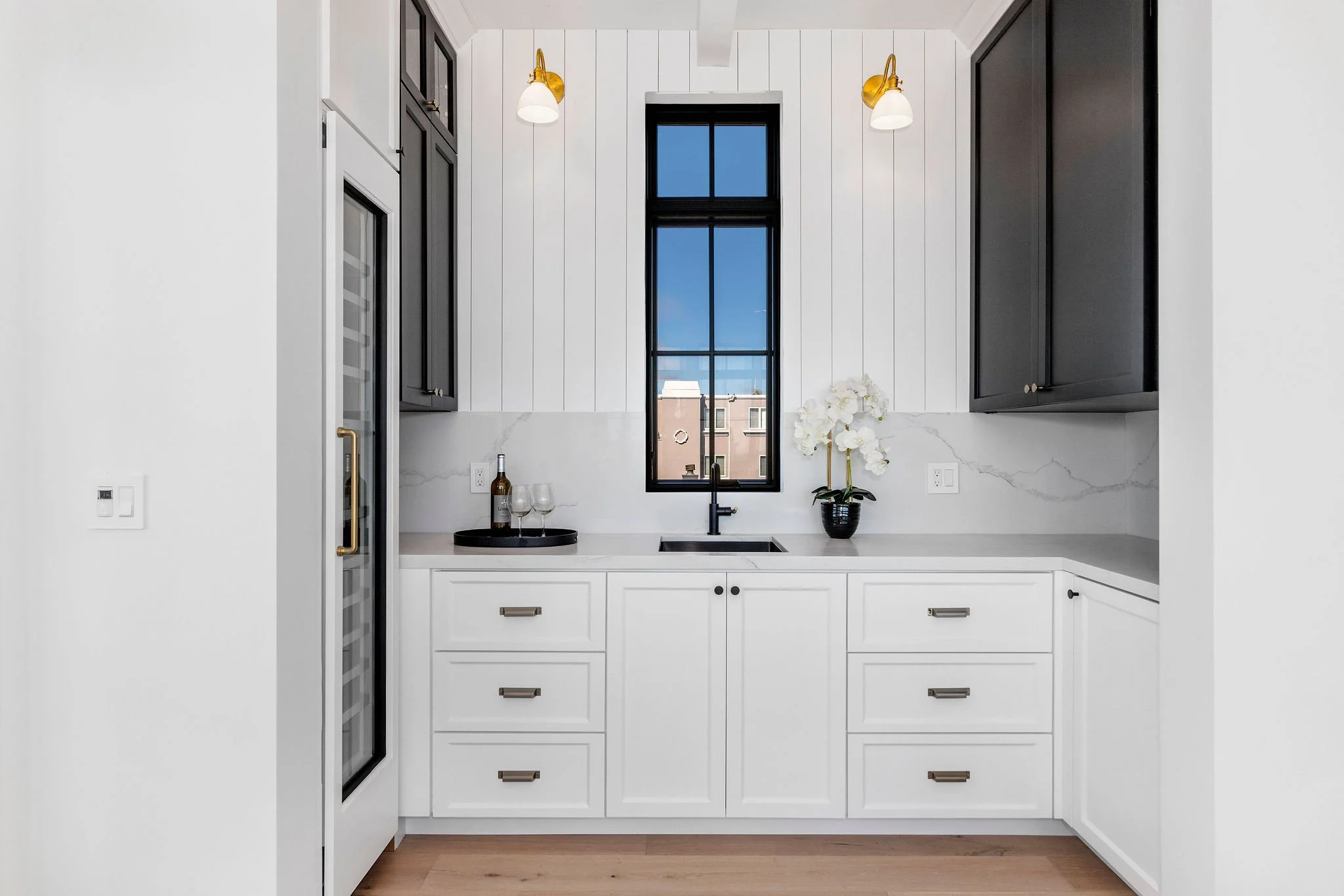 Modern kitchen with white cabinetry, black window, and marble countertops. Includes a wine fridge, sink, gold sconces, and decorative plant.