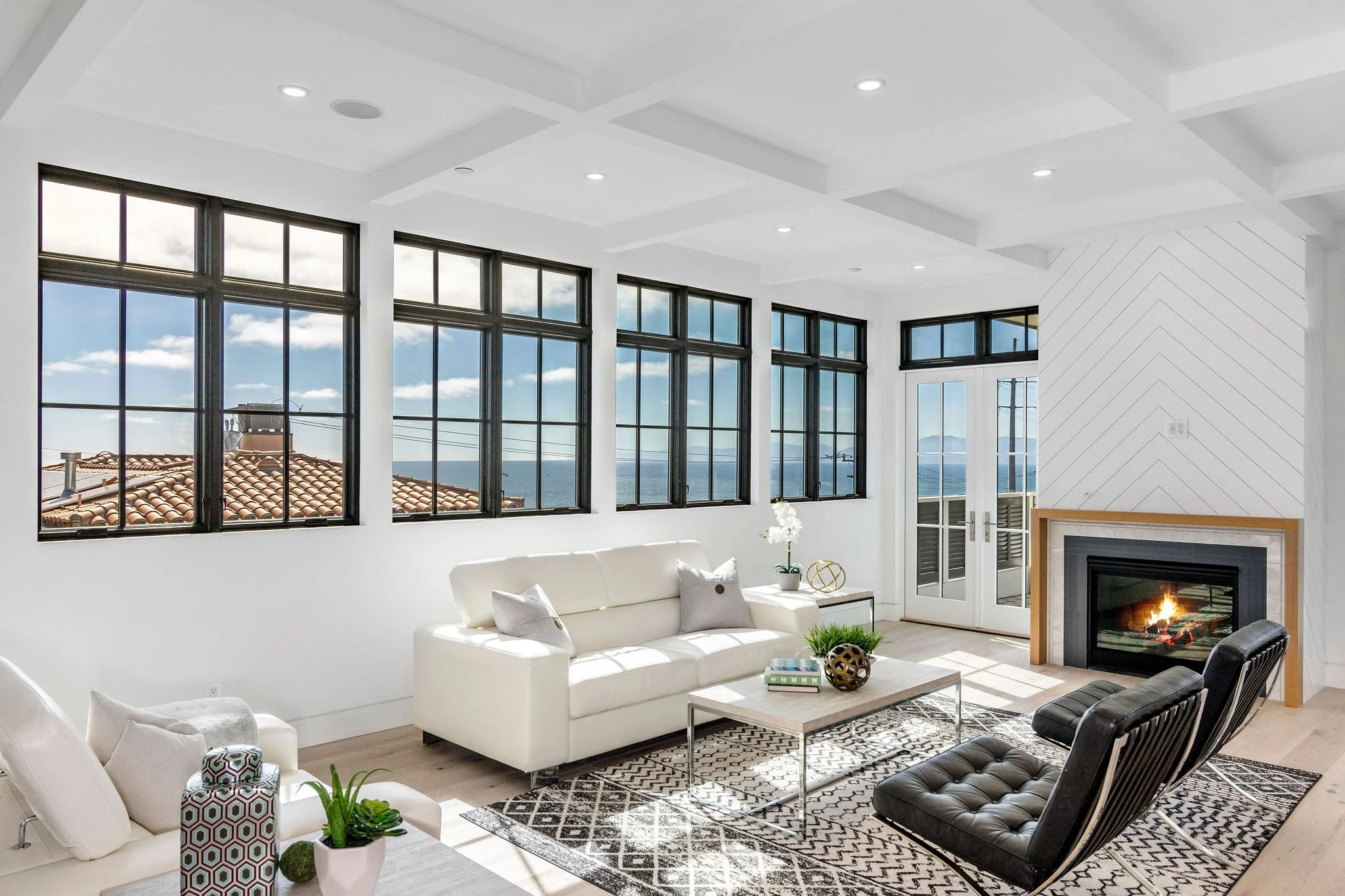 Modern living room with large windows, ocean view, white sofa, black chairs, fireplace, and geometric rug.