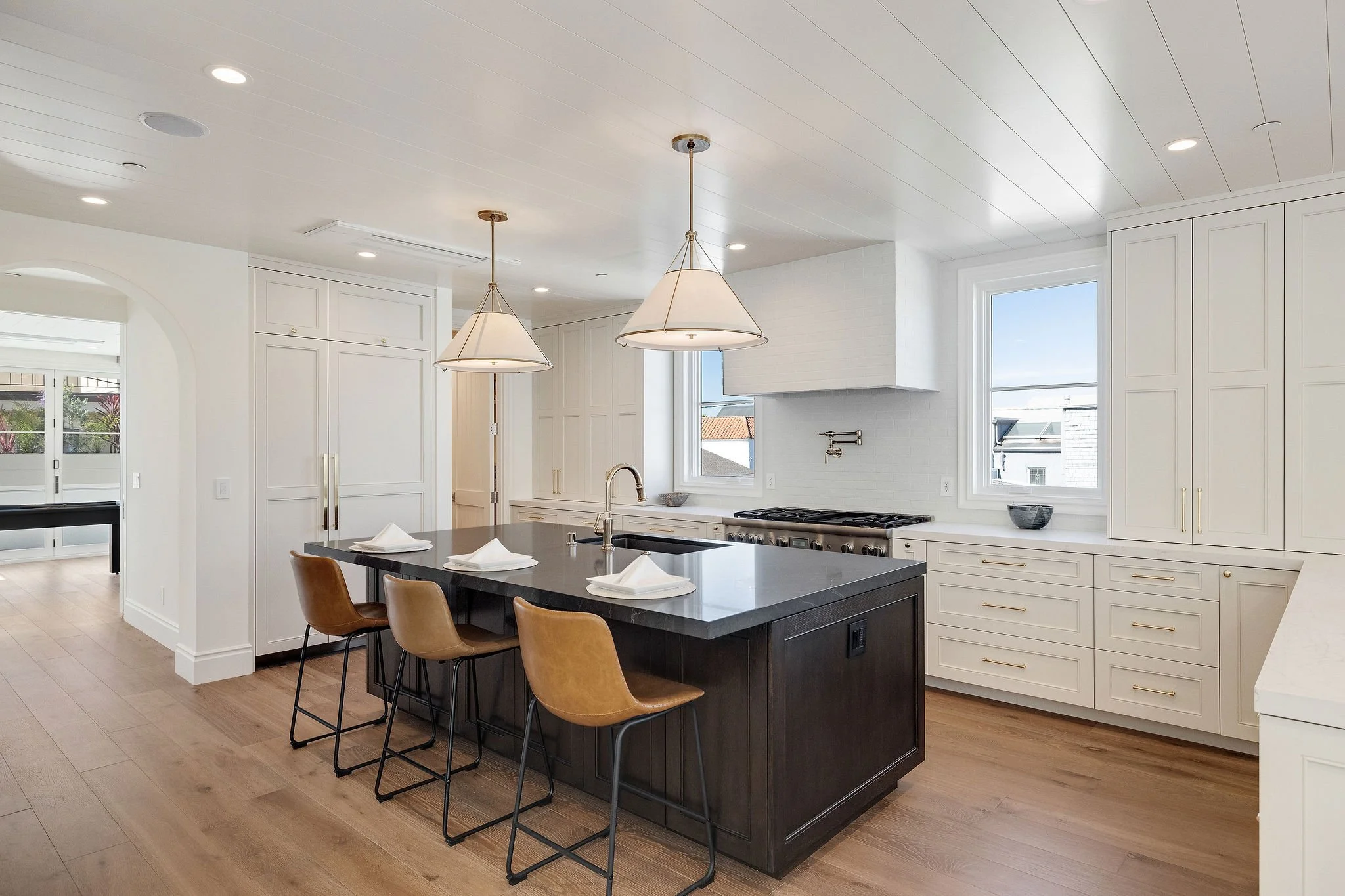Modern kitchen with an island, brown bar stools, pendant lights, white cabinets, and wooden flooring.