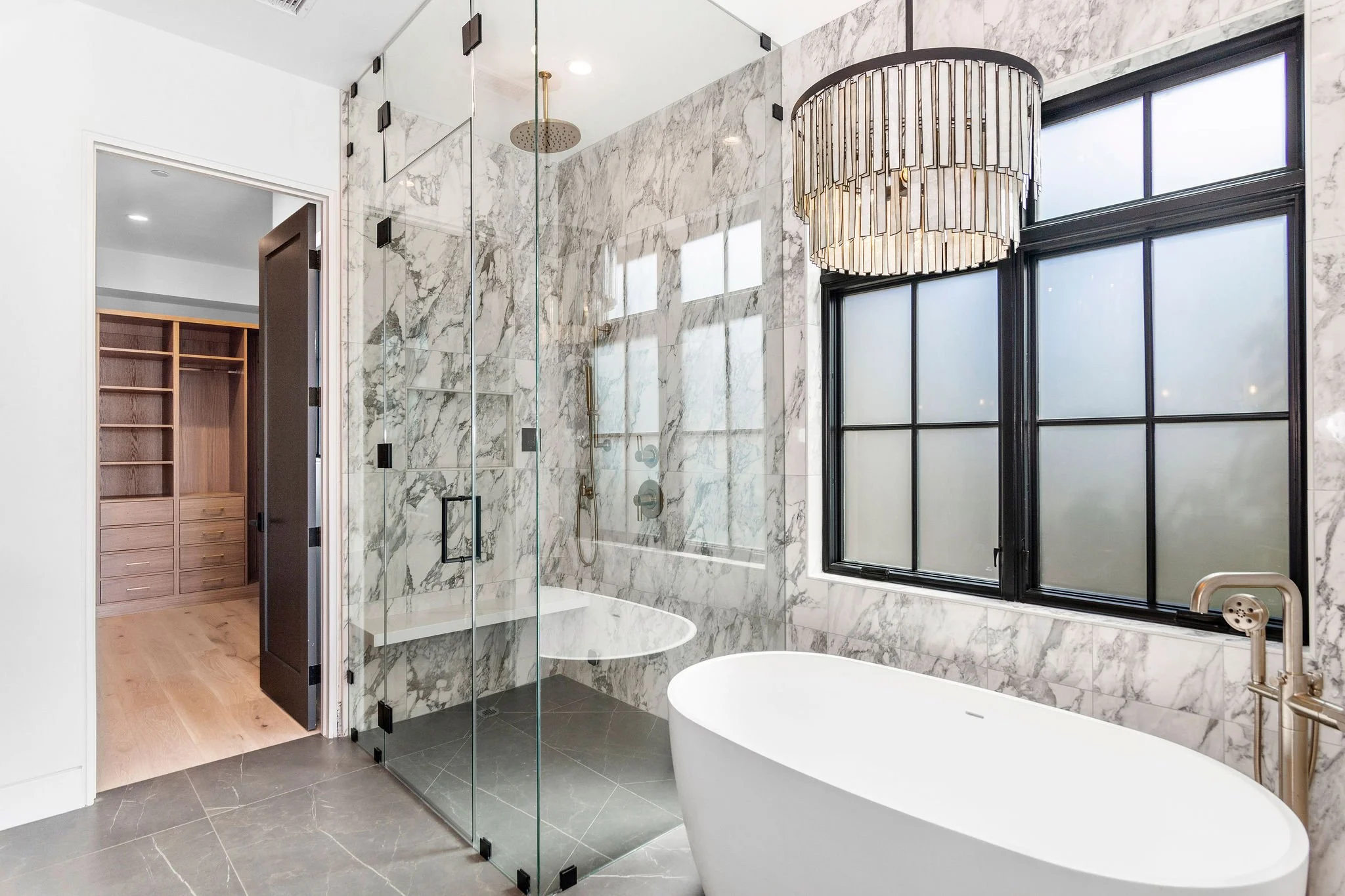 Modern bathroom with a freestanding bathtub, glass-enclosed shower, marble walls, large windows, and a chandelier. Visible walk-in closet through the open door.