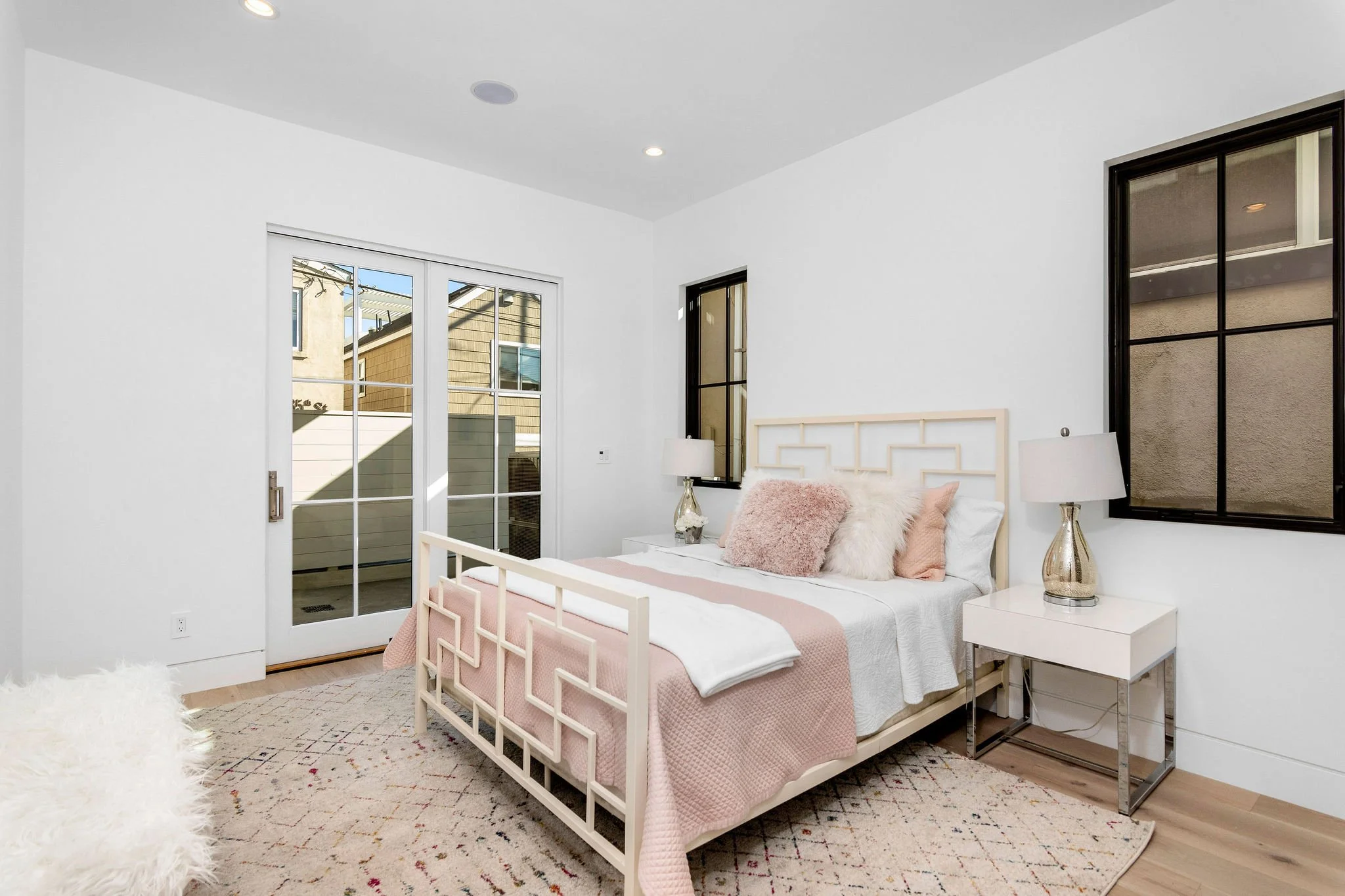 Modern bedroom with a bed featuring pink and white bedding, a patterned rug, nightstands with lamps, and large windows with a view outside.