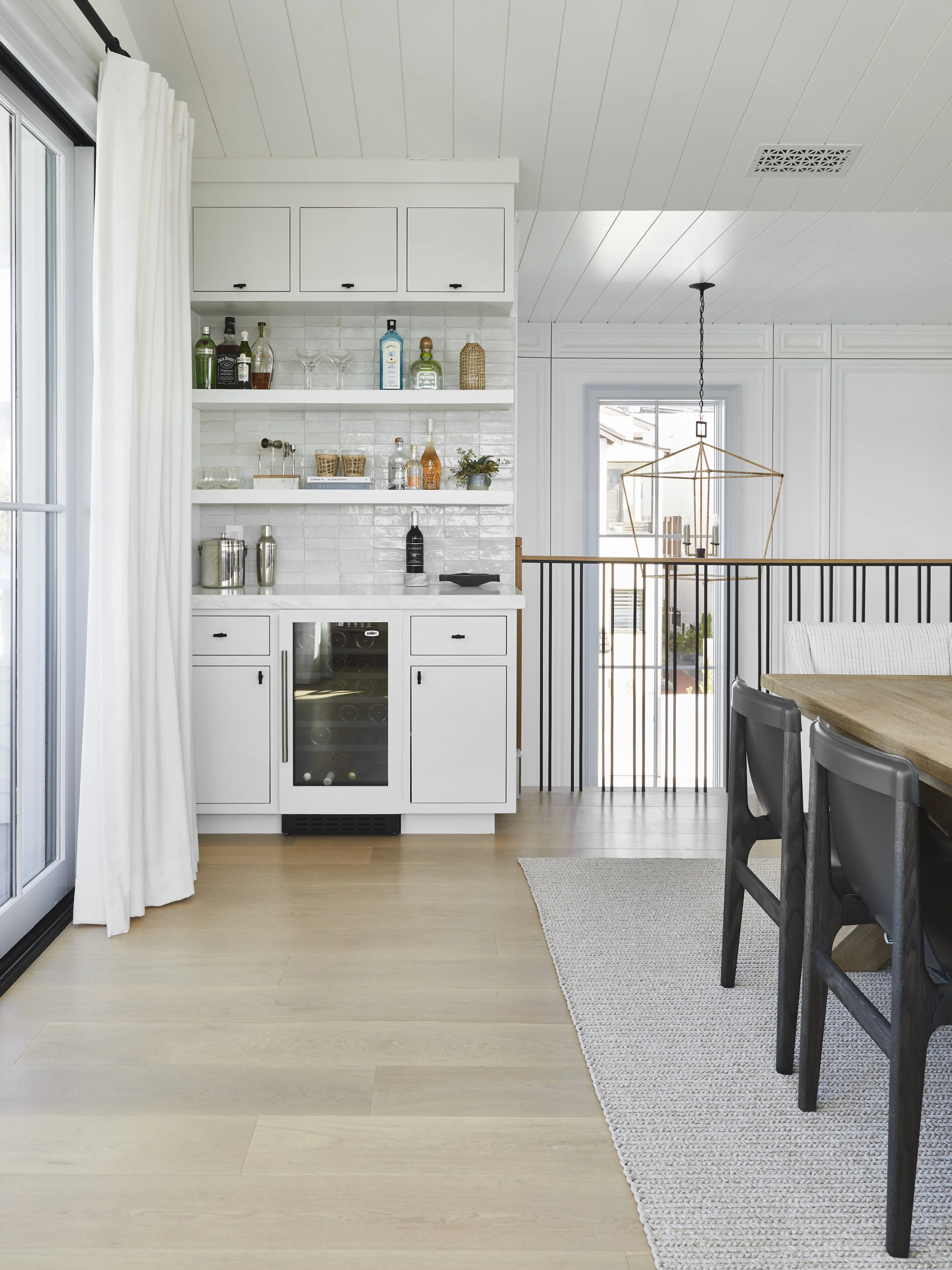 Modern home bar setup with white cabinetry, open shelves stocked with bottles and glassware, a small wine fridge, a wooden dining table with black chairs, and a white curtain on the left. The room has light wood flooring and a geometric pendant light