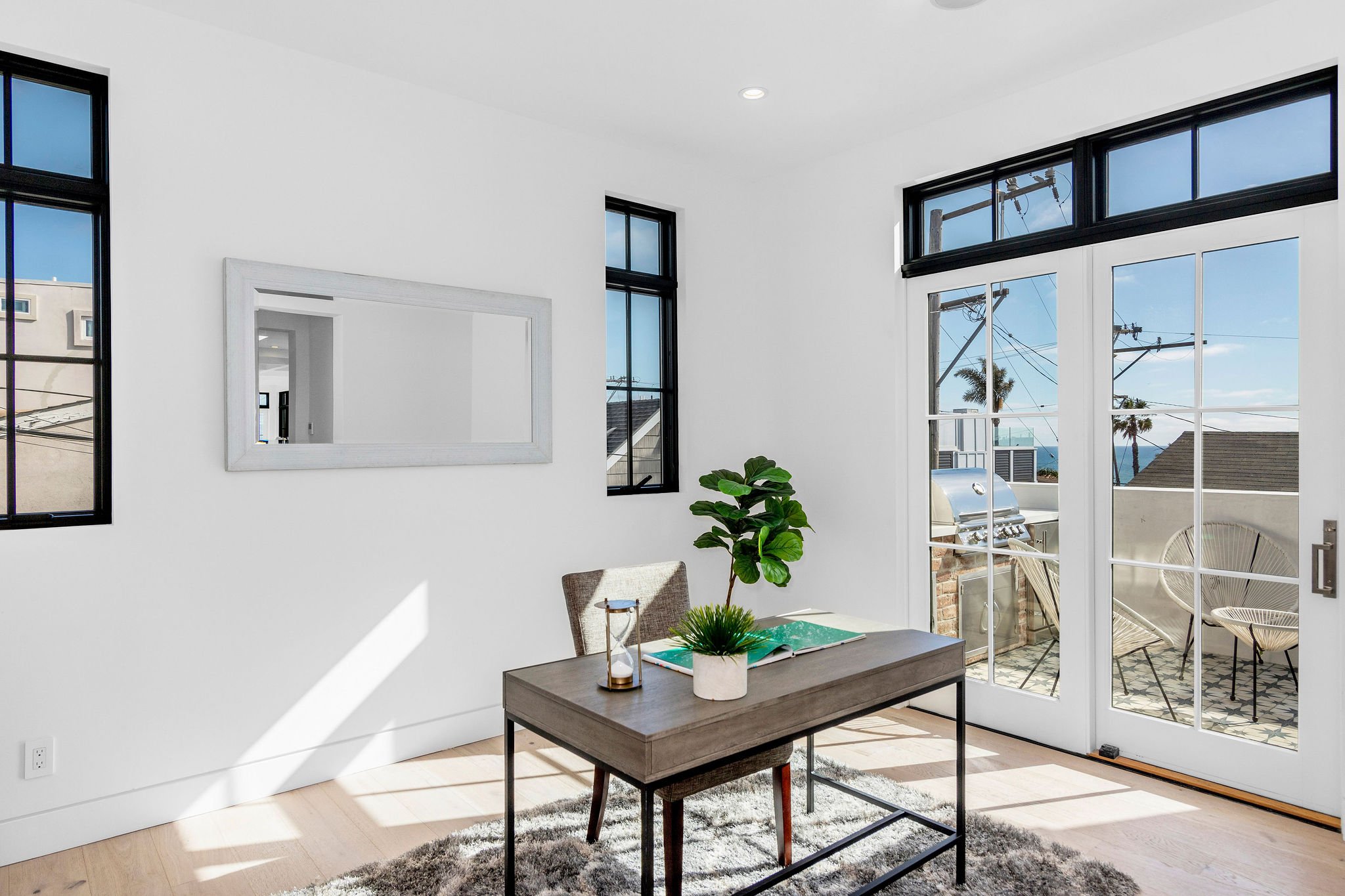 Bright, modern home office with a desk, chair, plants, large windows, and a view of a patio with outdoor furniture and a grill.