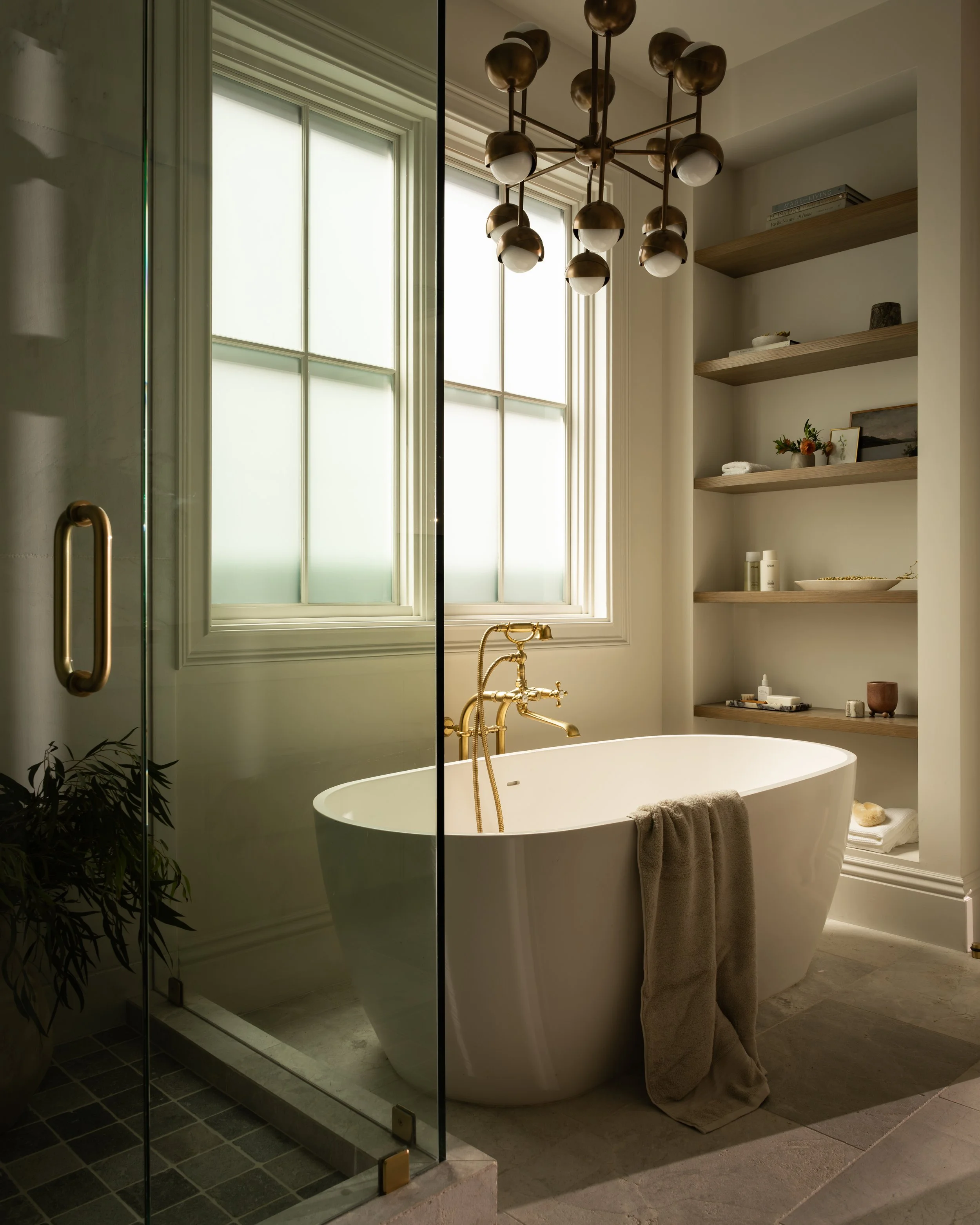 Modern bathroom with a freestanding white bathtub and gold fixtures, a hanging light fixture, built-in shelves with decor items, a towel draped over the tub, a glass shower door, and a plant in the corner.