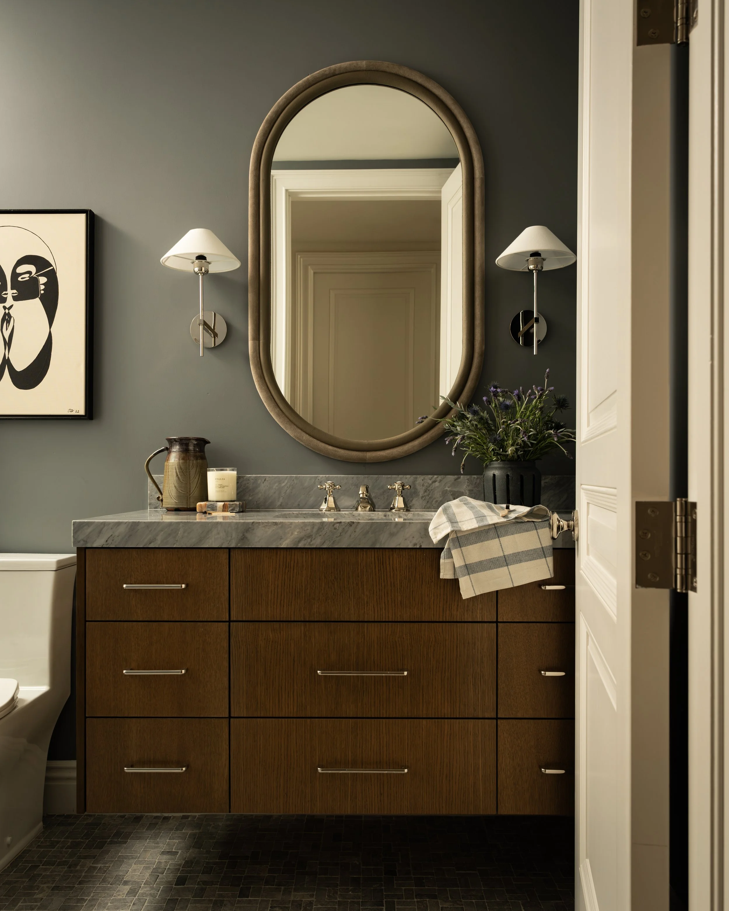 Modern bathroom interior with oval mirror, wooden vanity, marble countertop, wall sconces, abstract artwork, and decorative plants.