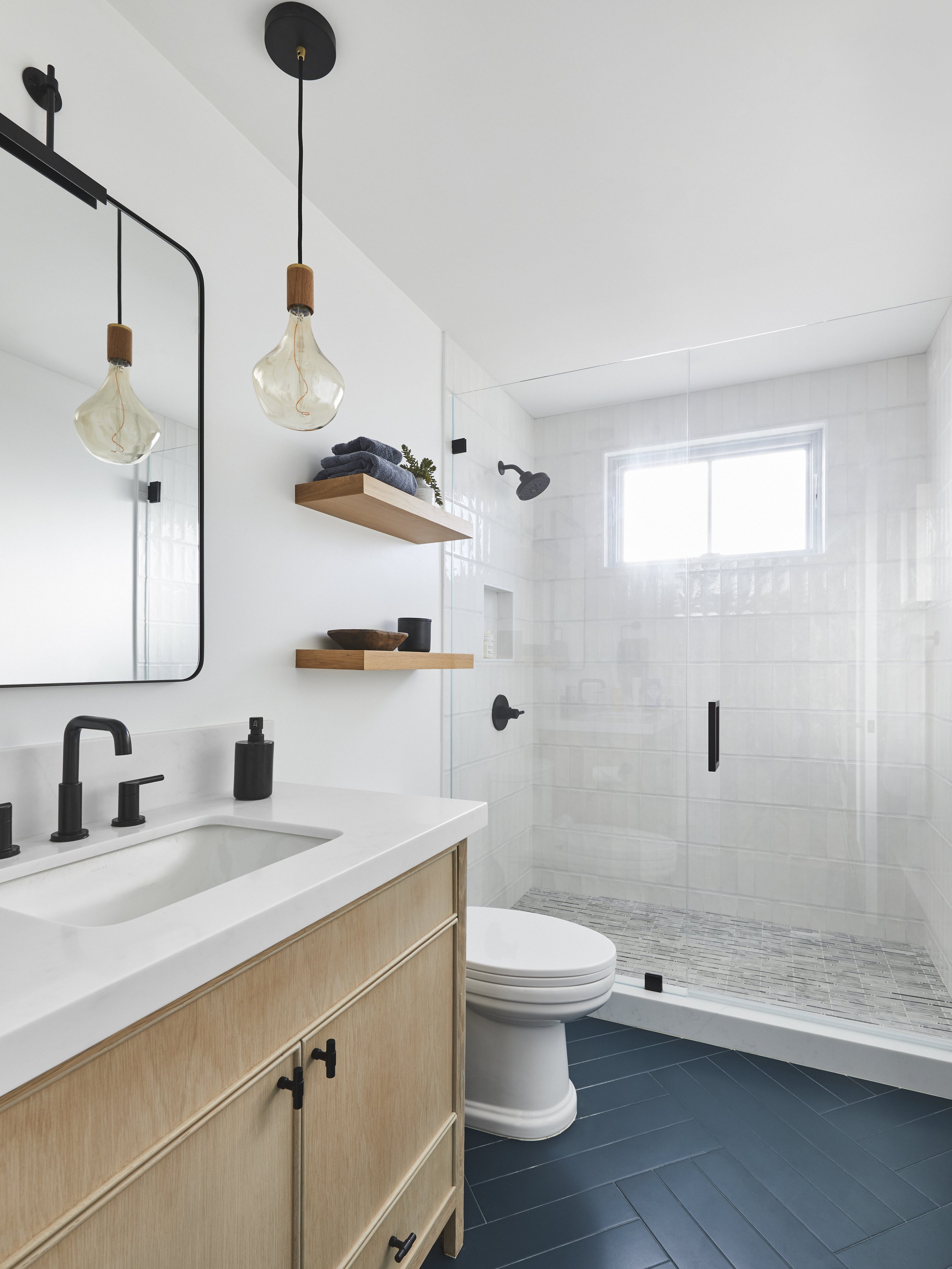 Modern bathroom with a wooden vanity and white countertop, black faucet, and a large mirror. Pendant lights hang above. The room features a toilet, open shelving with towels and decor, and a walk-in glass shower with black fixtures. Dark herringbone 