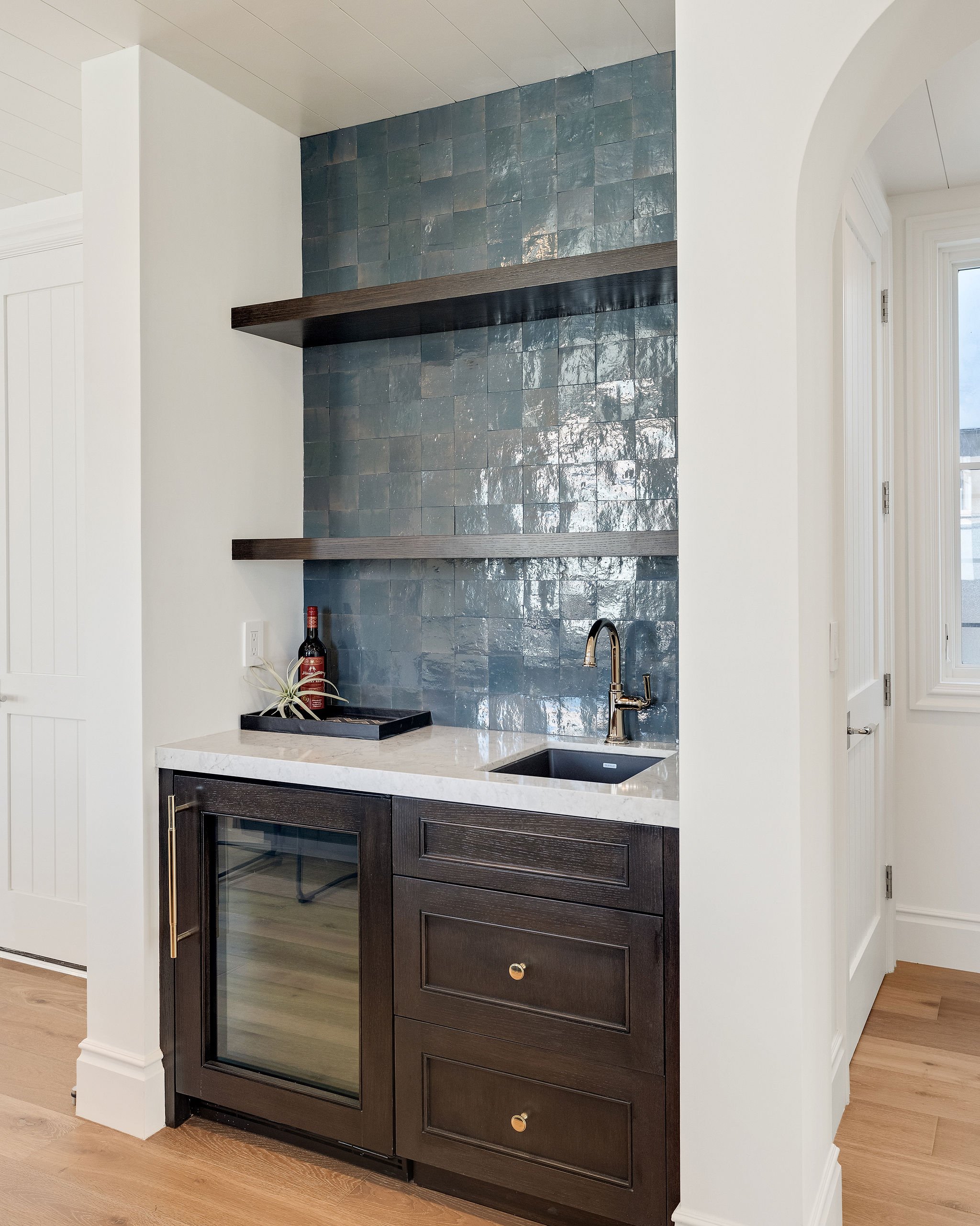 Small home bar with dark wood cabinetry, marble countertop, blue tile backsplash, open shelves, wine fridge, and faucet.
