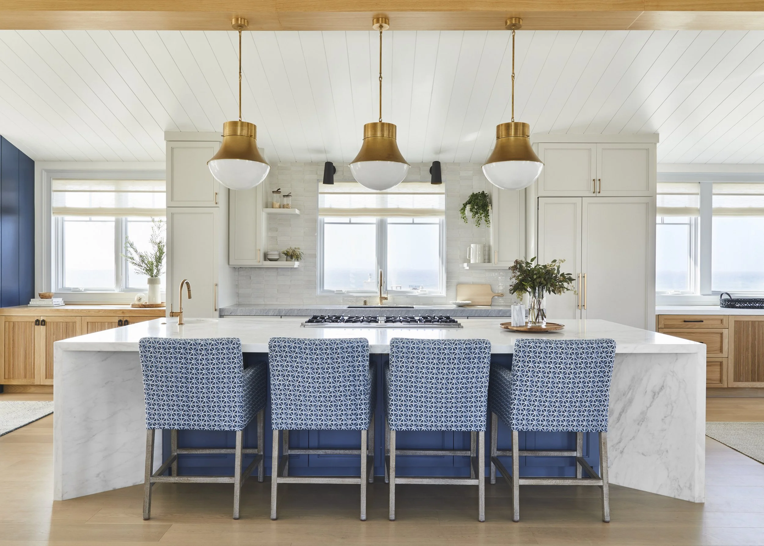 Modern kitchen with a large marble island, blue patterned bar stools, brass pendant lights, and light cabinetry.
