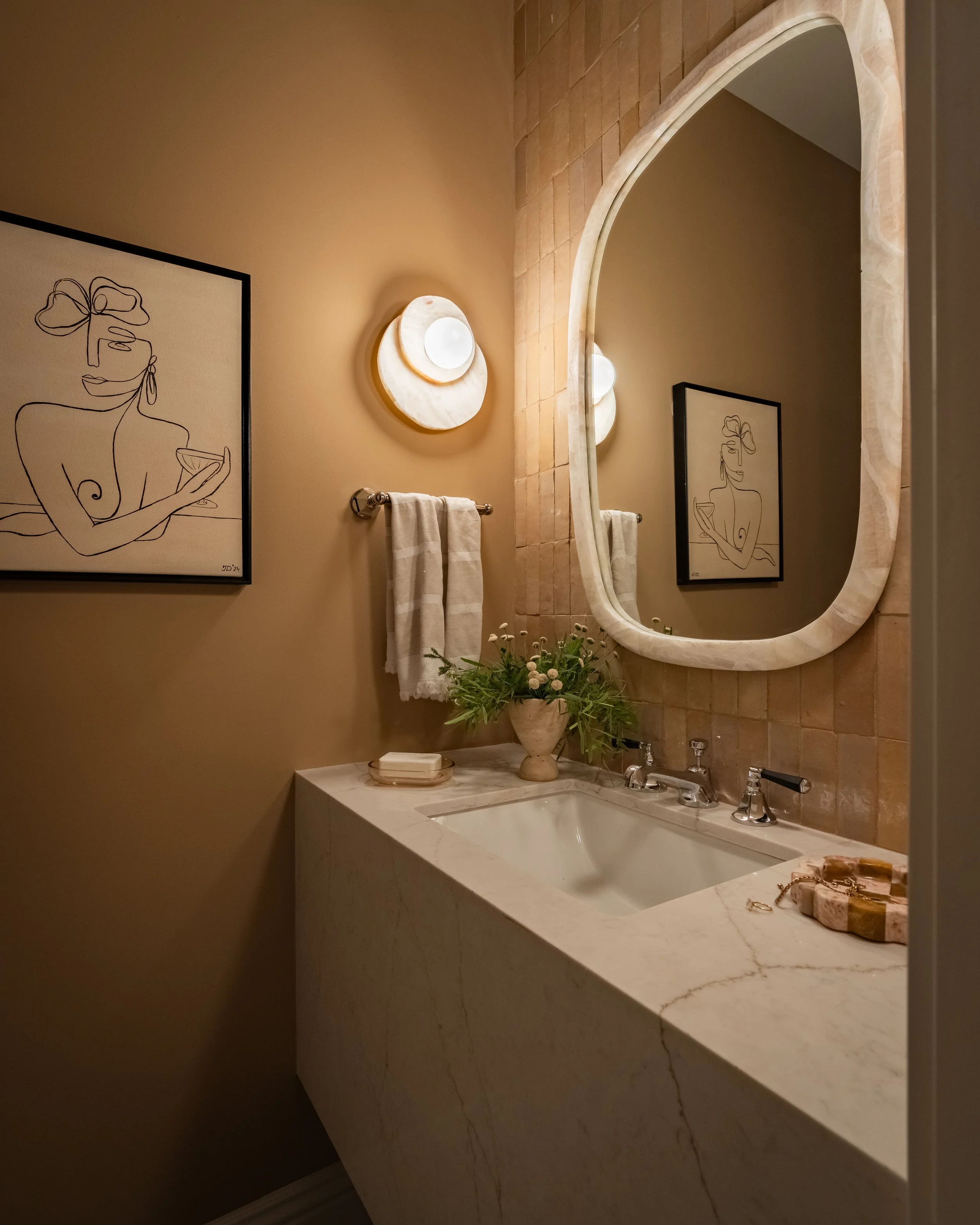 Bathroom with marble countertop, modern mirror, wall art, and round wall light.