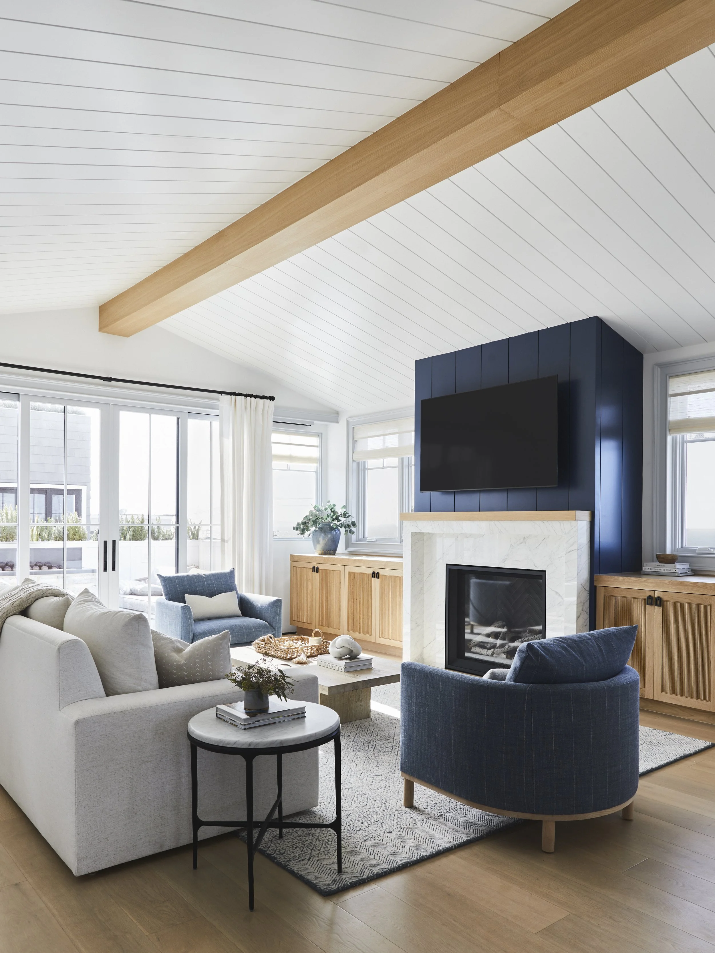 Modern living room with white walls, large windows, and wooden ceiling beam. Features a fireplace, cushioned sofa, armchairs, marble side table, and light wood flooring.