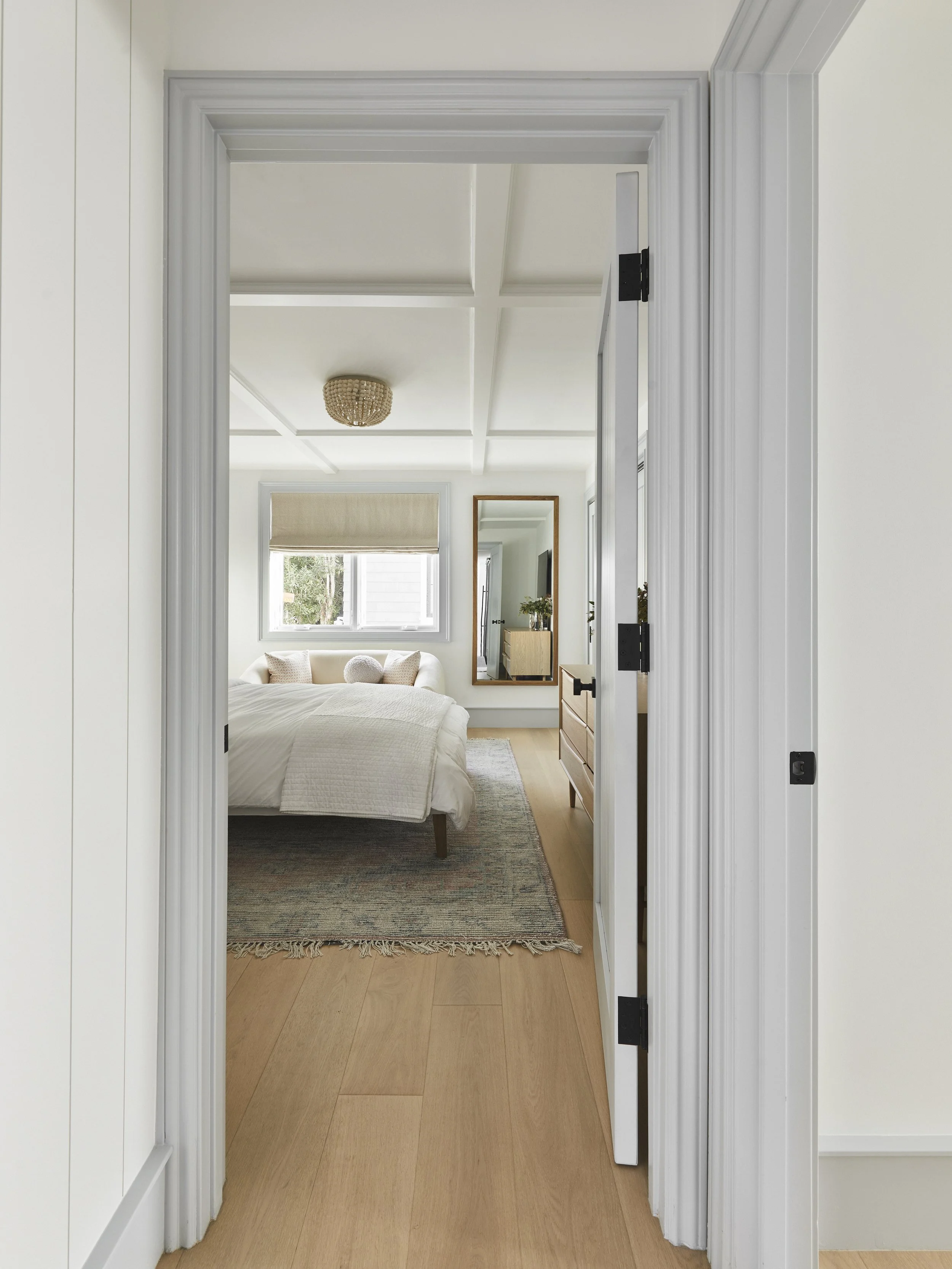 View through a doorway into a modern bedroom with a white bed, light wood flooring, and a woven light fixture. A large mirror and dresser are visible, along with a window with a beige blind.